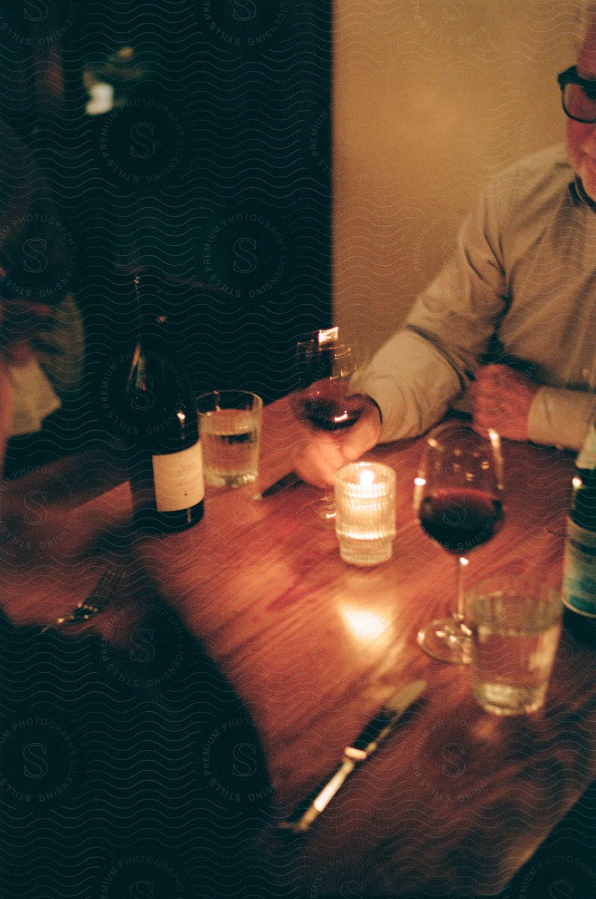 Dinner setting with wine glasses and a bottle on a wooden table, illuminated by candlelight, with a person in the background.