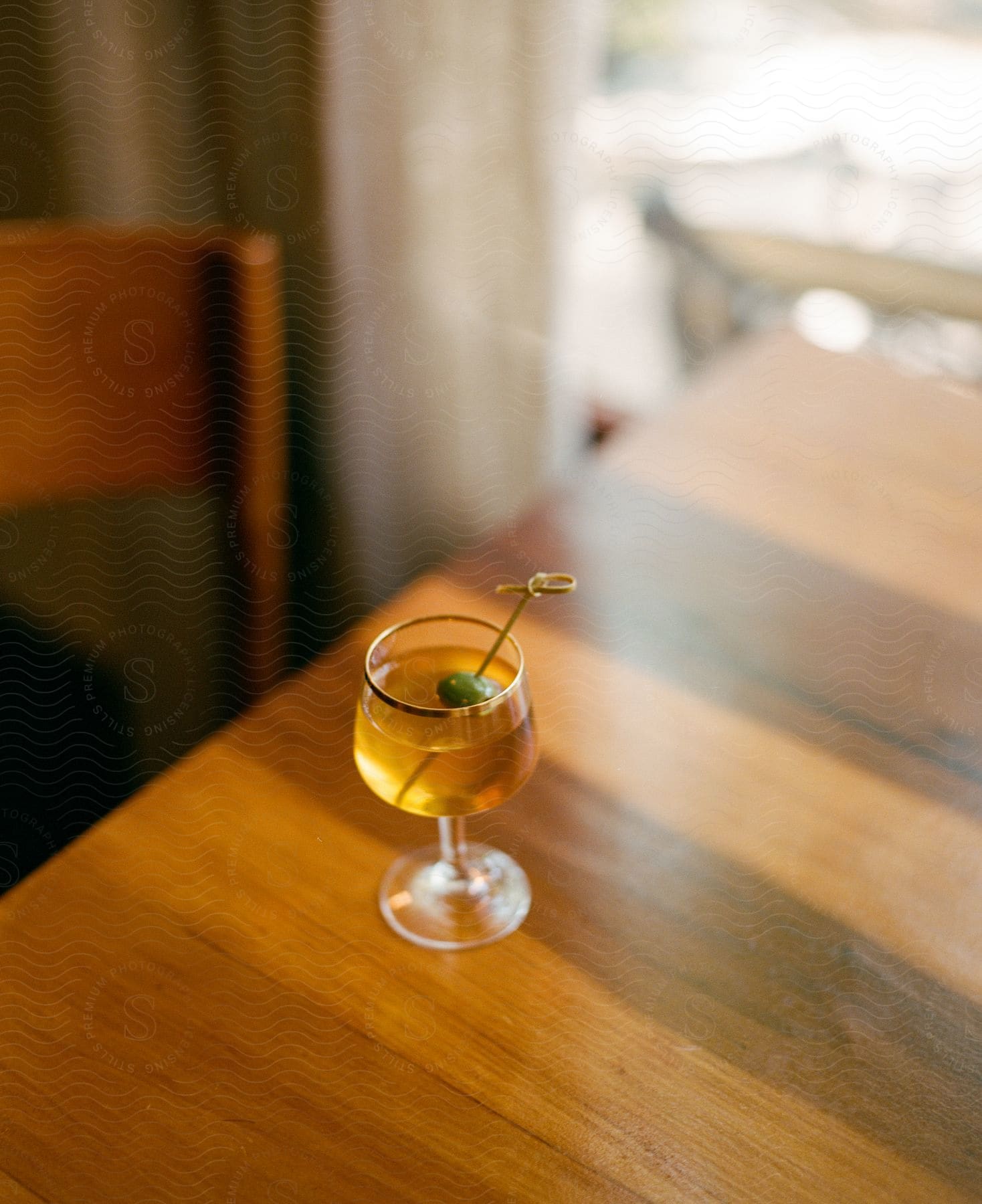 A drink with an olive sits on a table by a window