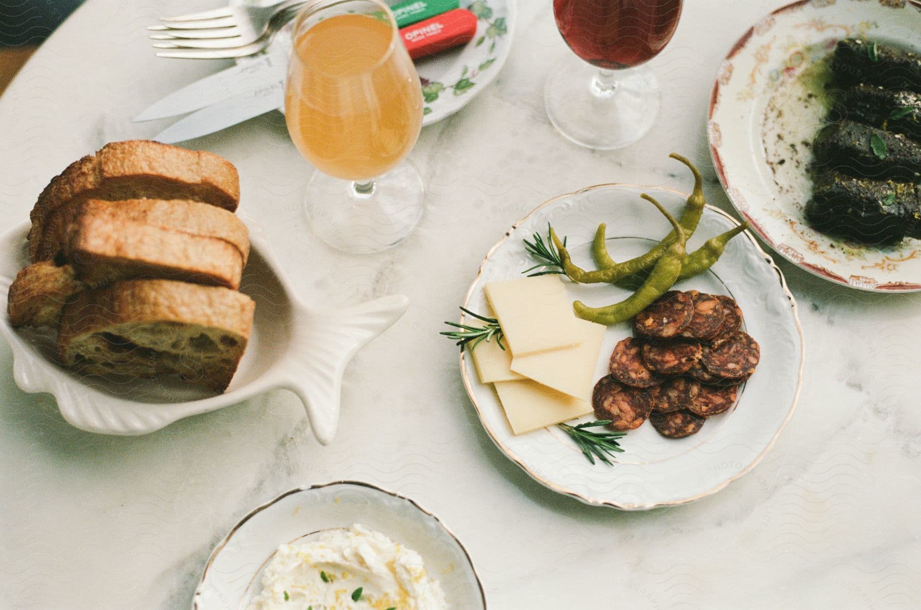 Assorted appetizers on a marble surface including sliced bread, green pickled peppers, chorizo slices, tzatziki sauce, and beverages in stemware.