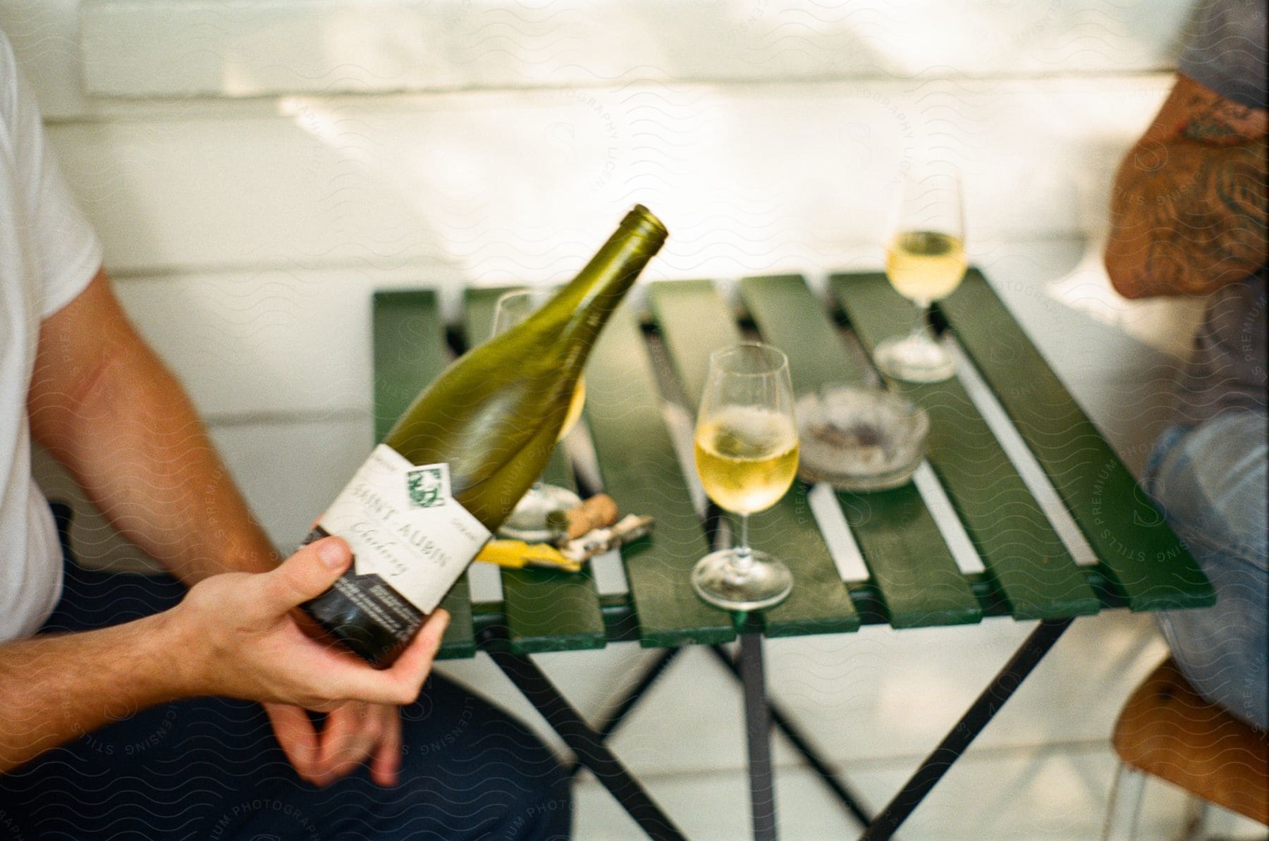 A person holding a wine bottle over a green slatted table with three wine glasses and an ash tray.