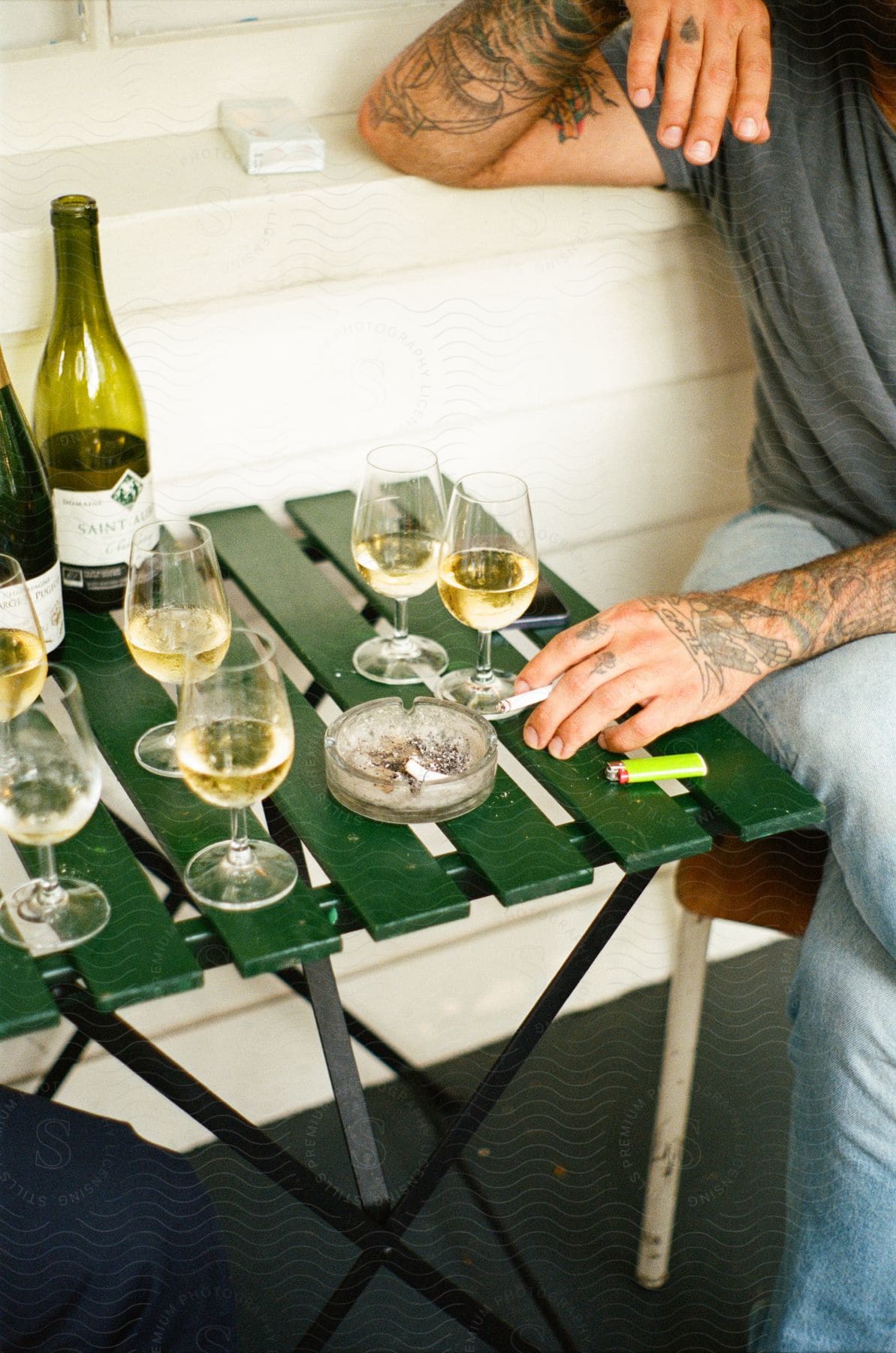 A man with tattoos smoking at a small table which is covered in wine glasses and a couple of bottles