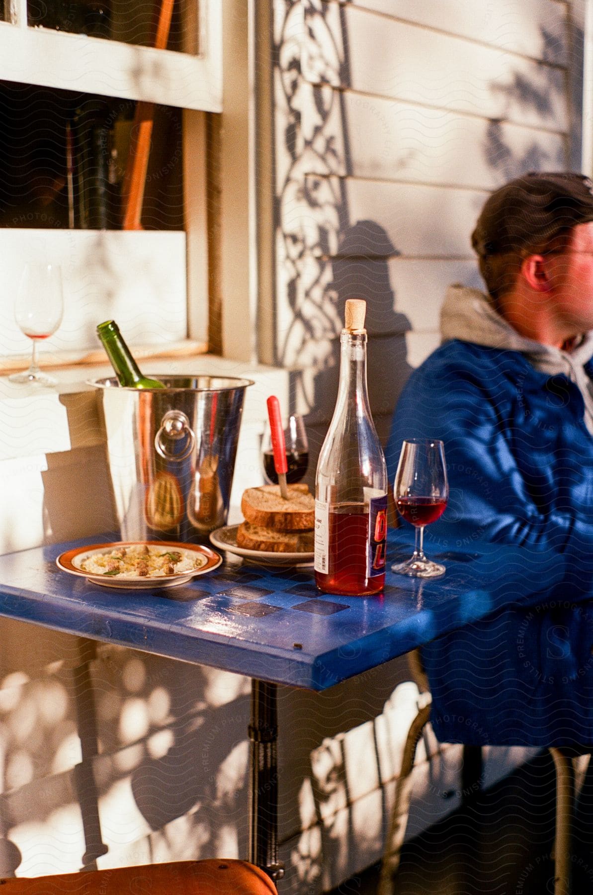 A sandwich, wine glasses and bottles of alcohol on a blue checkerboard table next to a man in a blue coat.