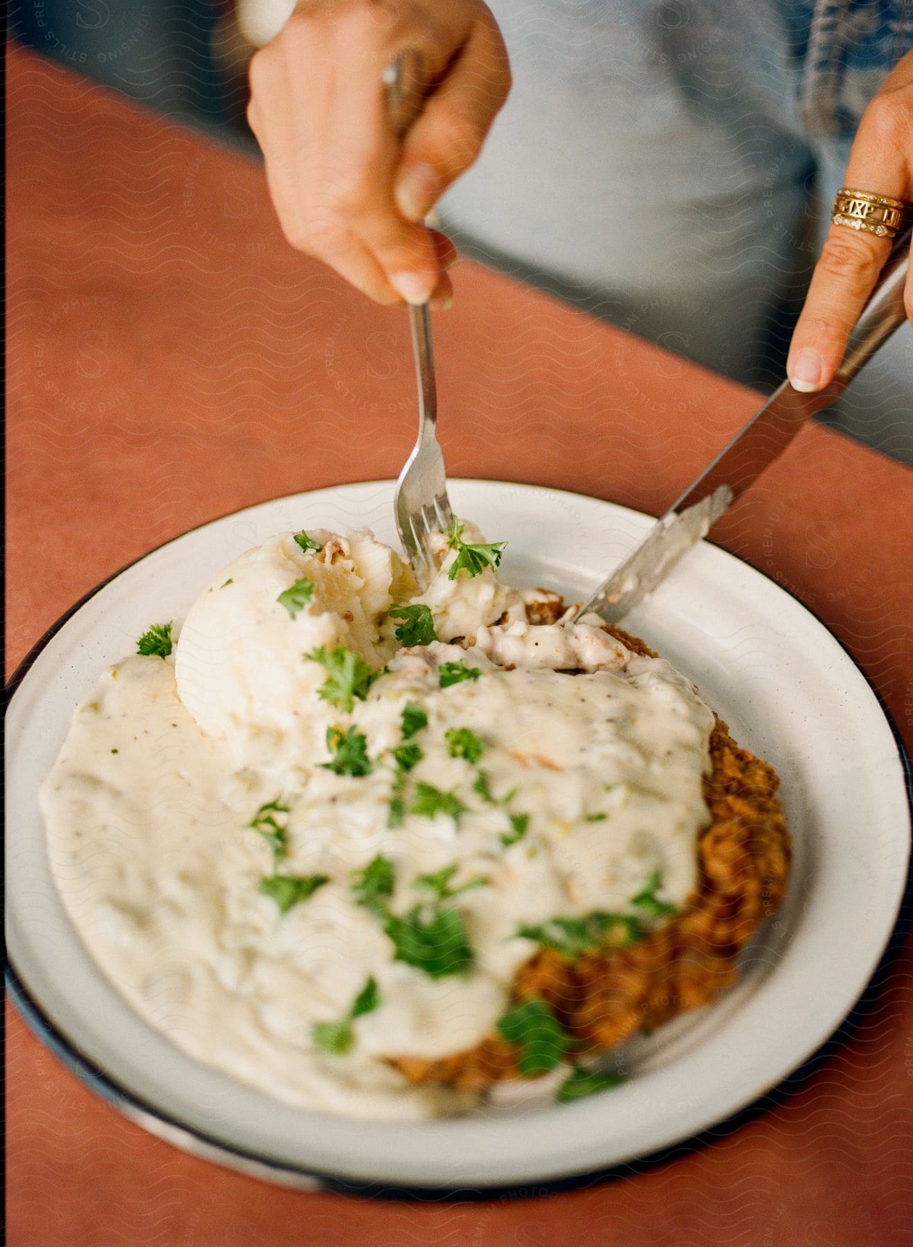 Human hands holding a fork and a knife that are on a plate of food on a table and the plate has a white sauce and full of green vegetables