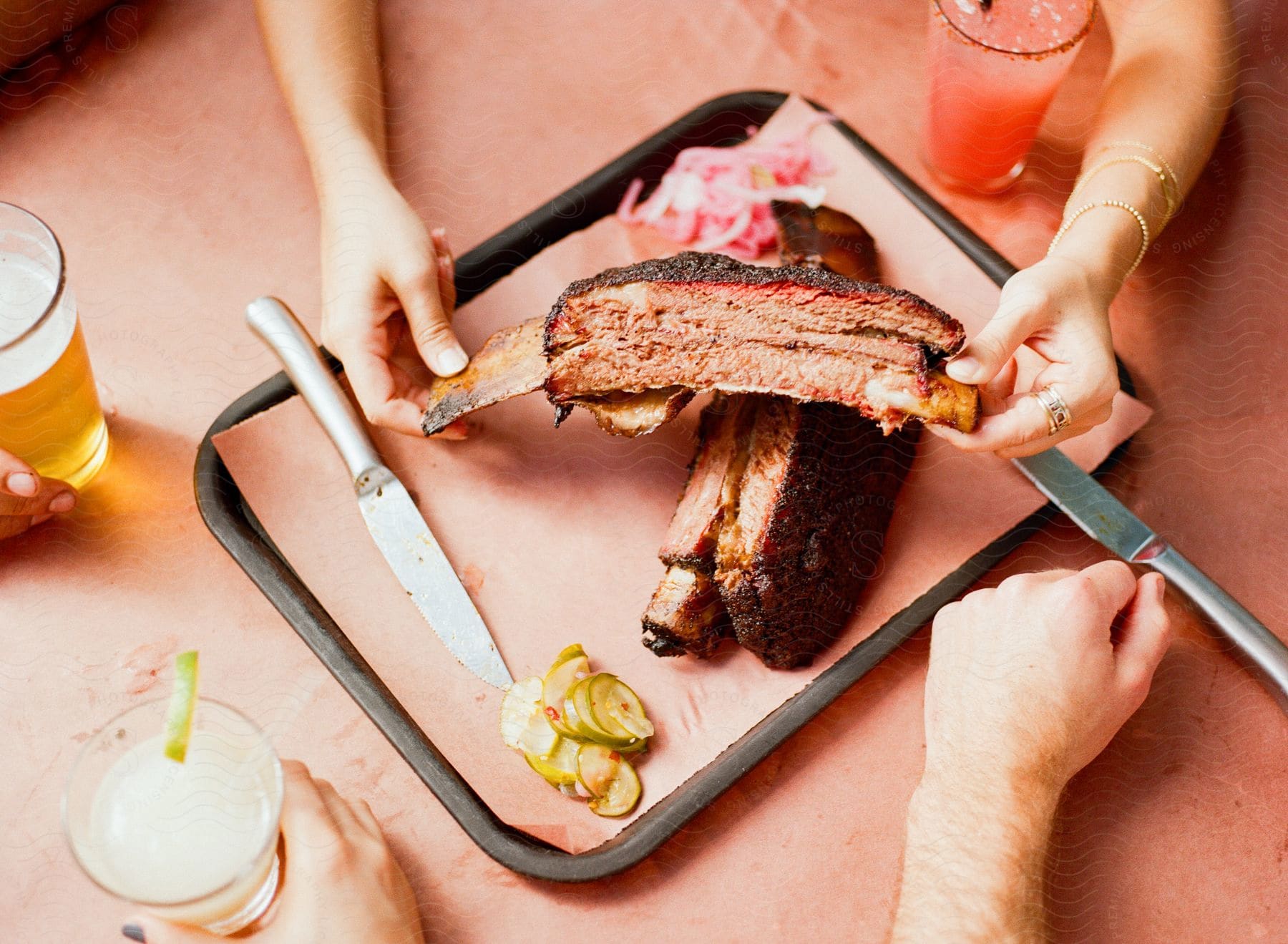 People sharing a large barbecued rib on a tray, with pickles, drinks, and condiments on the side.