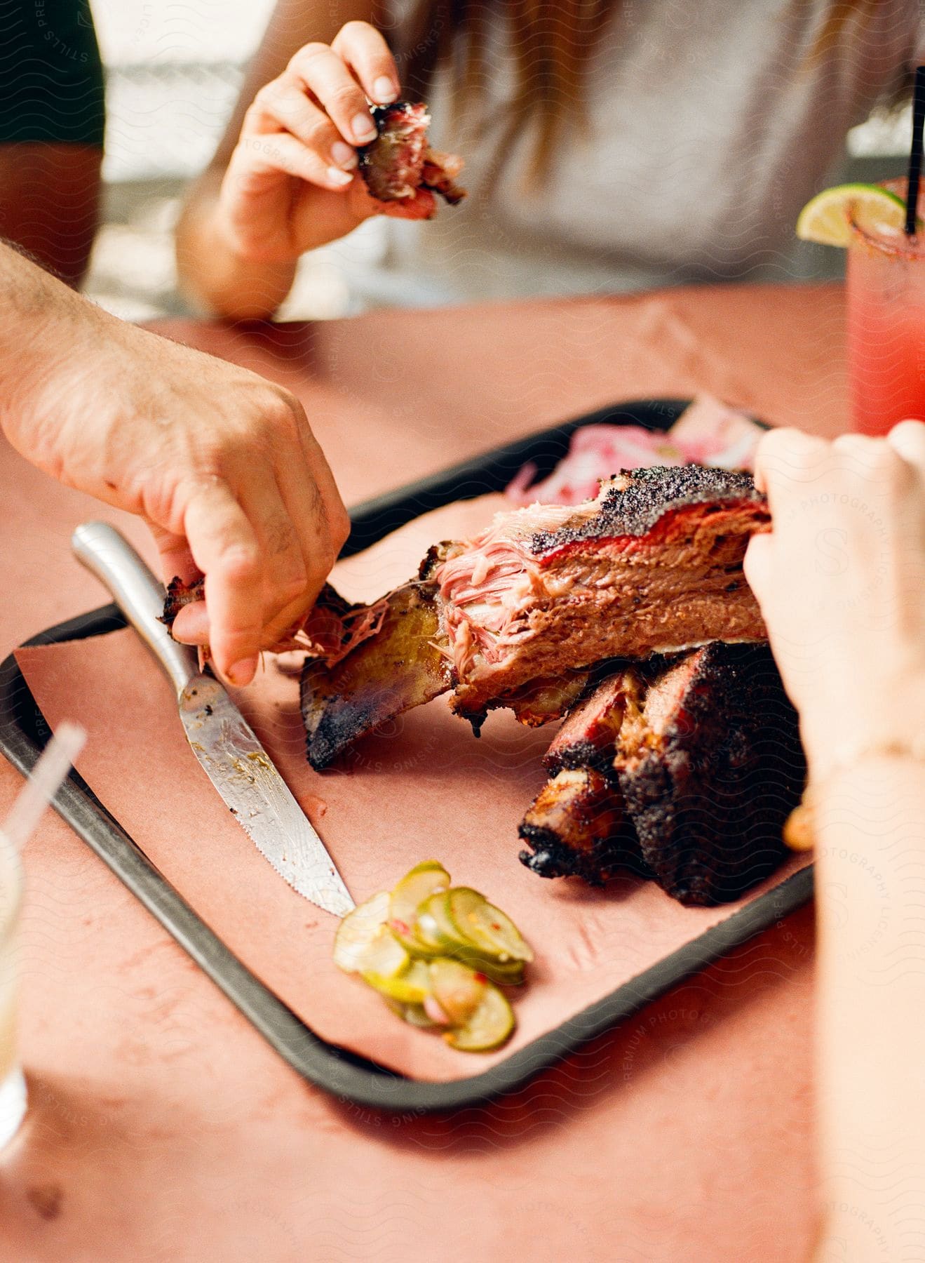 Stock photo of a woman is sitting at a table eating ribs as a man reaches for a rib on a tray with sliced pickles