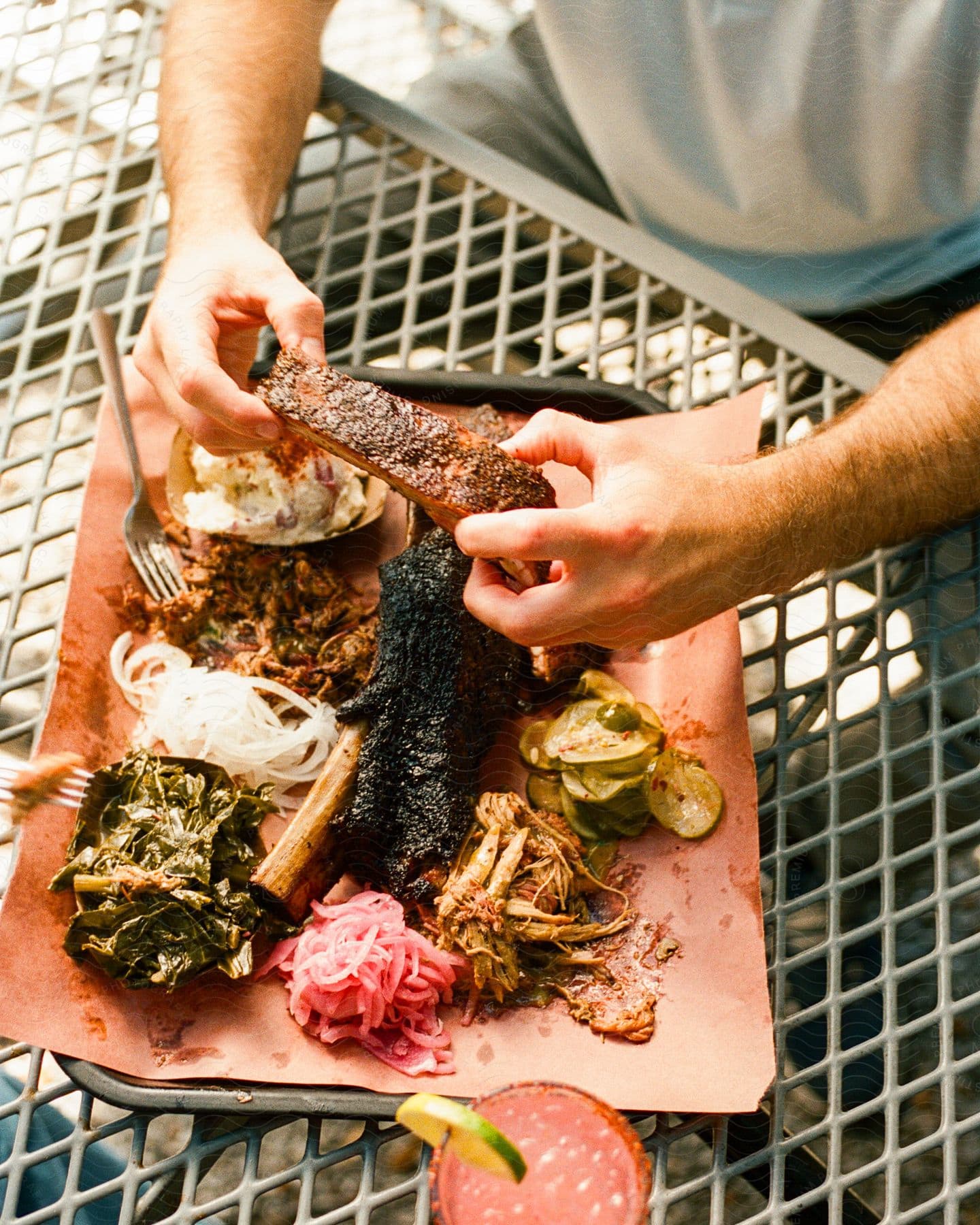 Person holding a barbecued rib over a paper-lined tray of assorted smoked meats and sides on a metal table.
