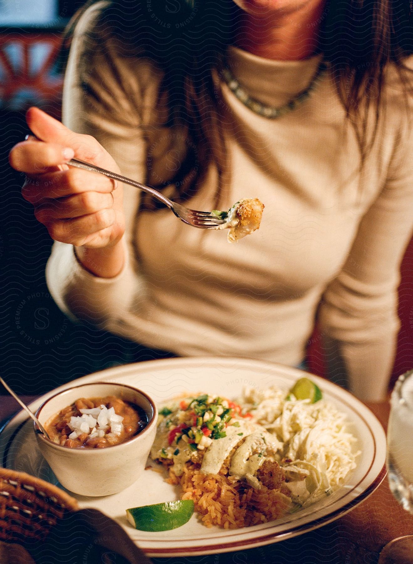 Woman eating a piece of food on a fork with a plate of Mexican cuisine in the background, featuring rice, beans, and enchiladas.