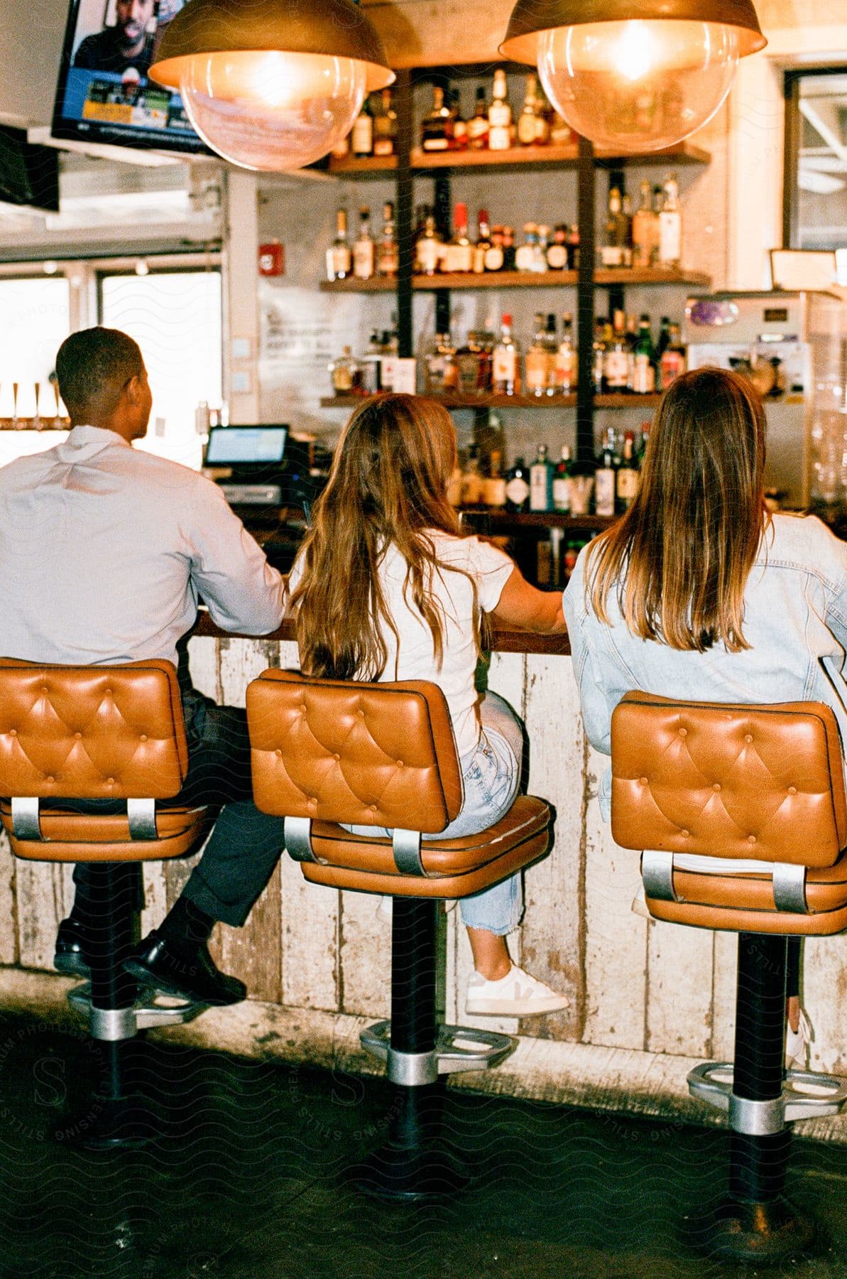 Two women and a man are sitting at a bar with bottles of alcohol on the shelves in front of them.