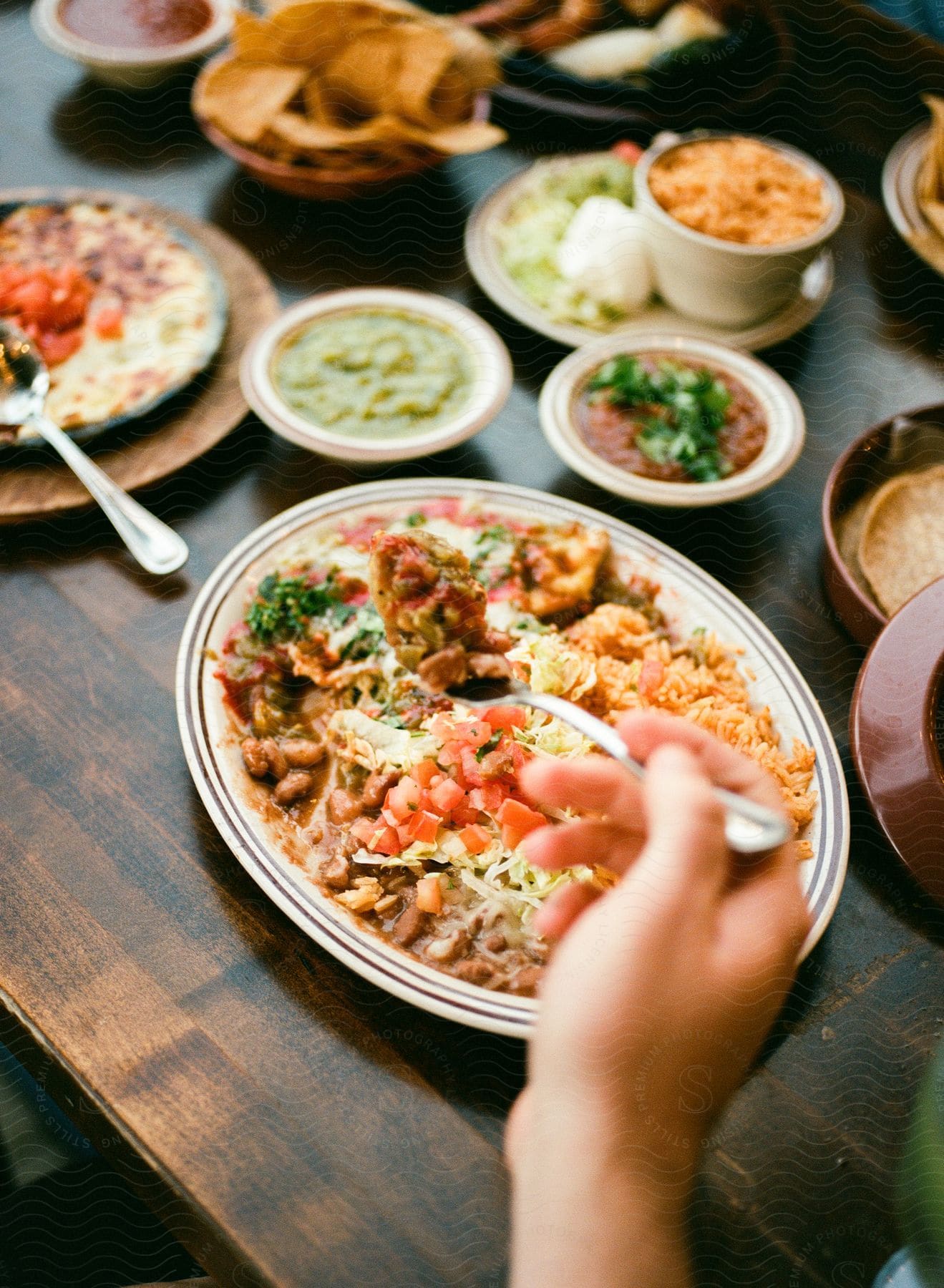 A hand holding a forkful from an enchilada plate with a table full of other mexican dishes