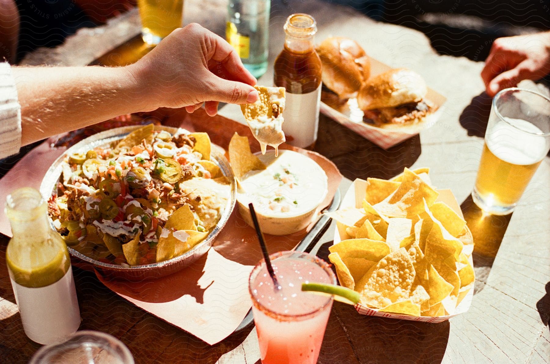 Person dipping a tortilla chip into a bowl of queso beside a plate of loaded nachos, with beverages on a sunny table.