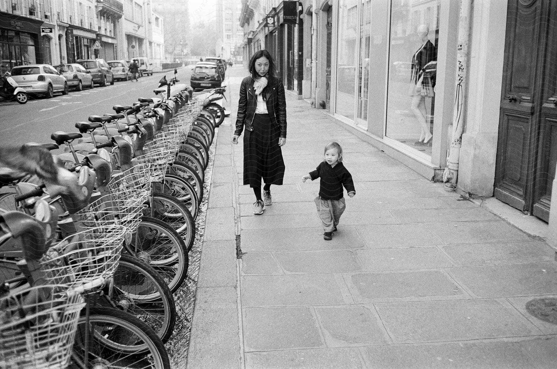 A woman strolling down a deserted street, accompanied by a toddler on the sidewalk, surrounded by a row of parked bicycles along the roadside.