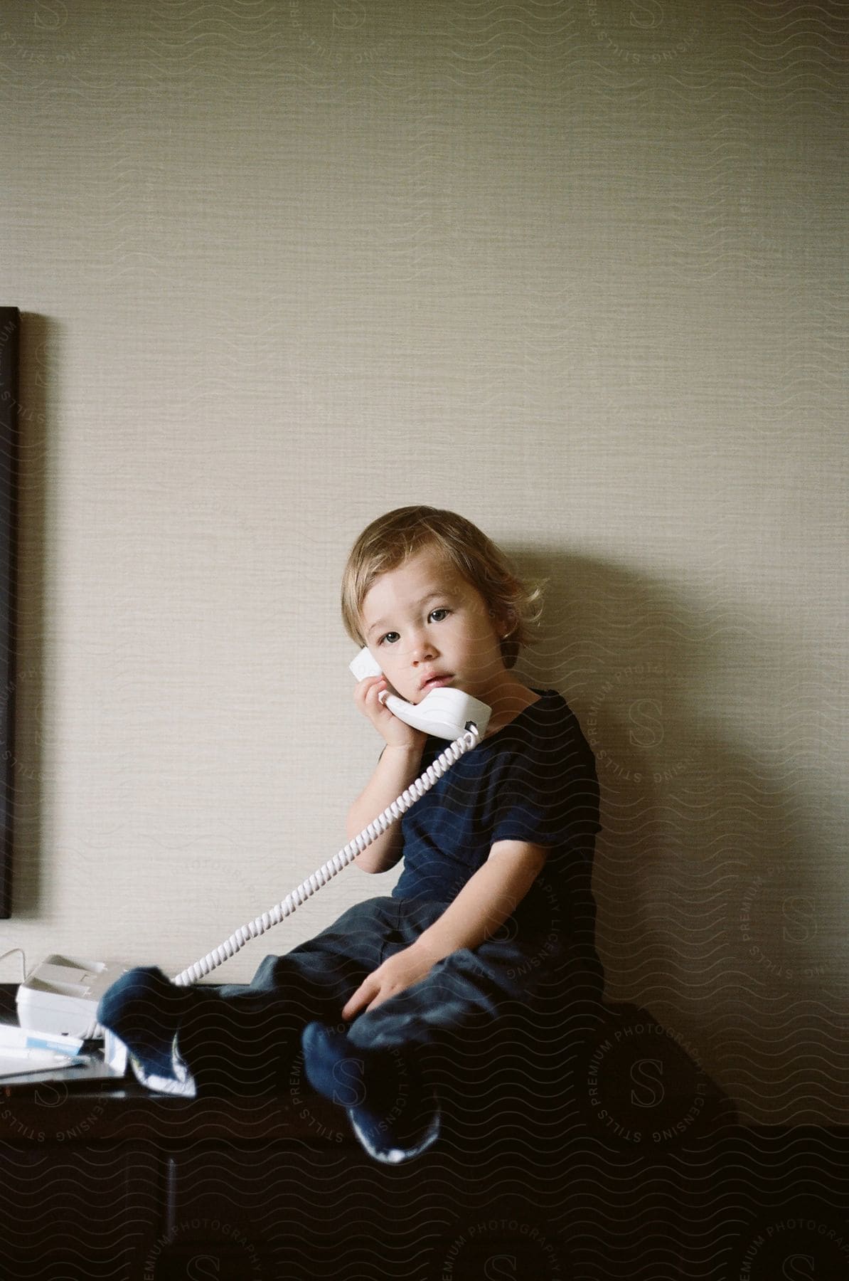 A blonde toddler sitting on a countertop with a corded telephone to her ear.