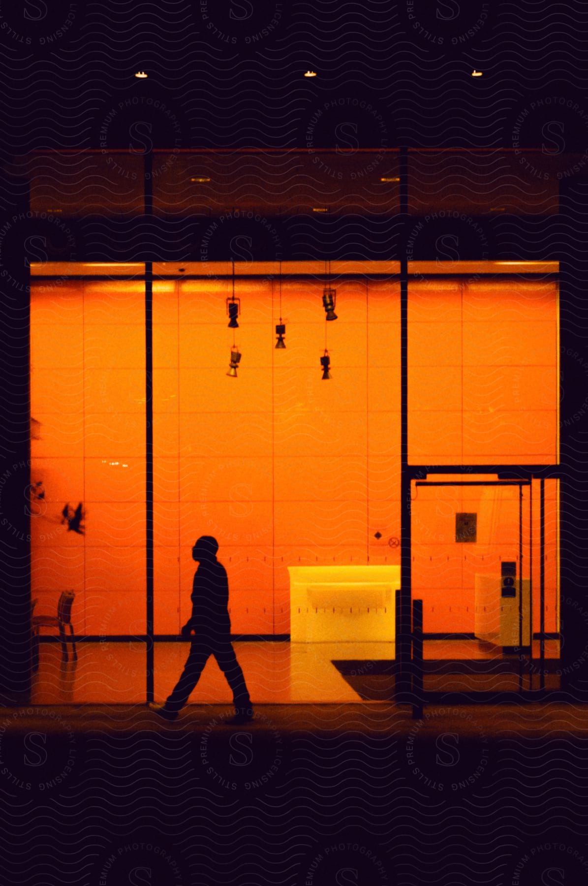 Silhouette of a man walking beside an illuminated hallway with warm lights at night.