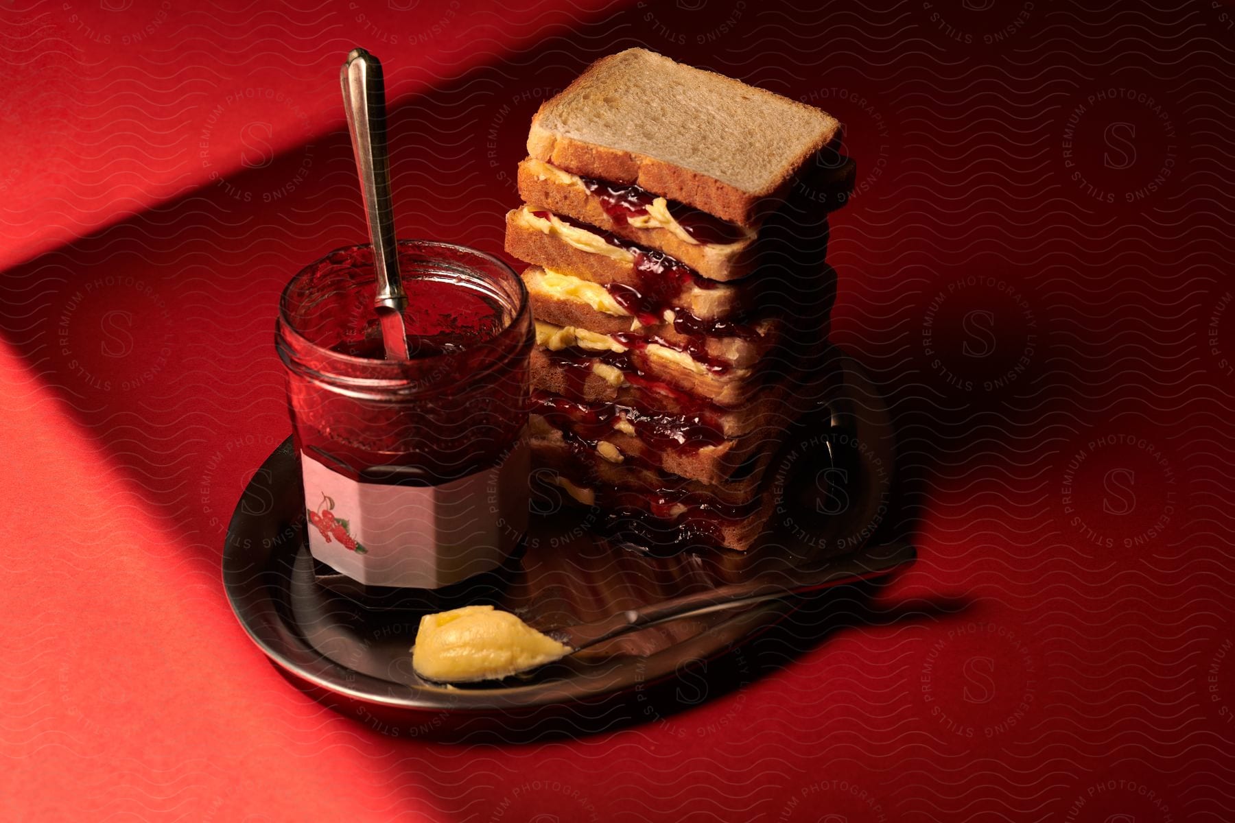 Peanut butter and jelly sandwiches are stacked on a tray with a jar of jelly and a knife with peanut butter