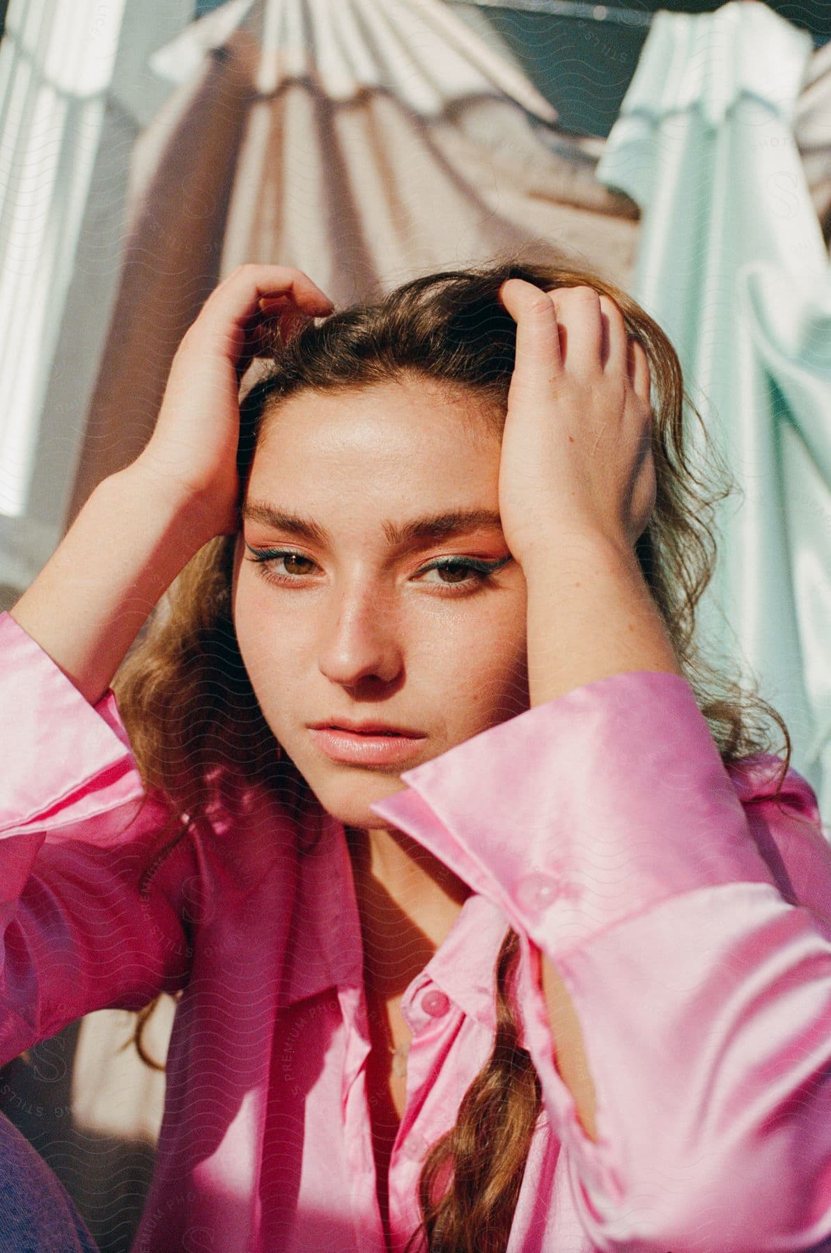 A woman wearing a silky pink blouse posed with both hands on her head in front of a fabric backdrop