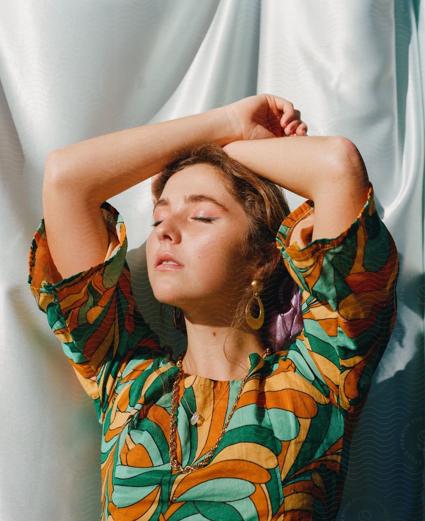 Fashion model crosses her wrists on her head near a fabric backdrop.