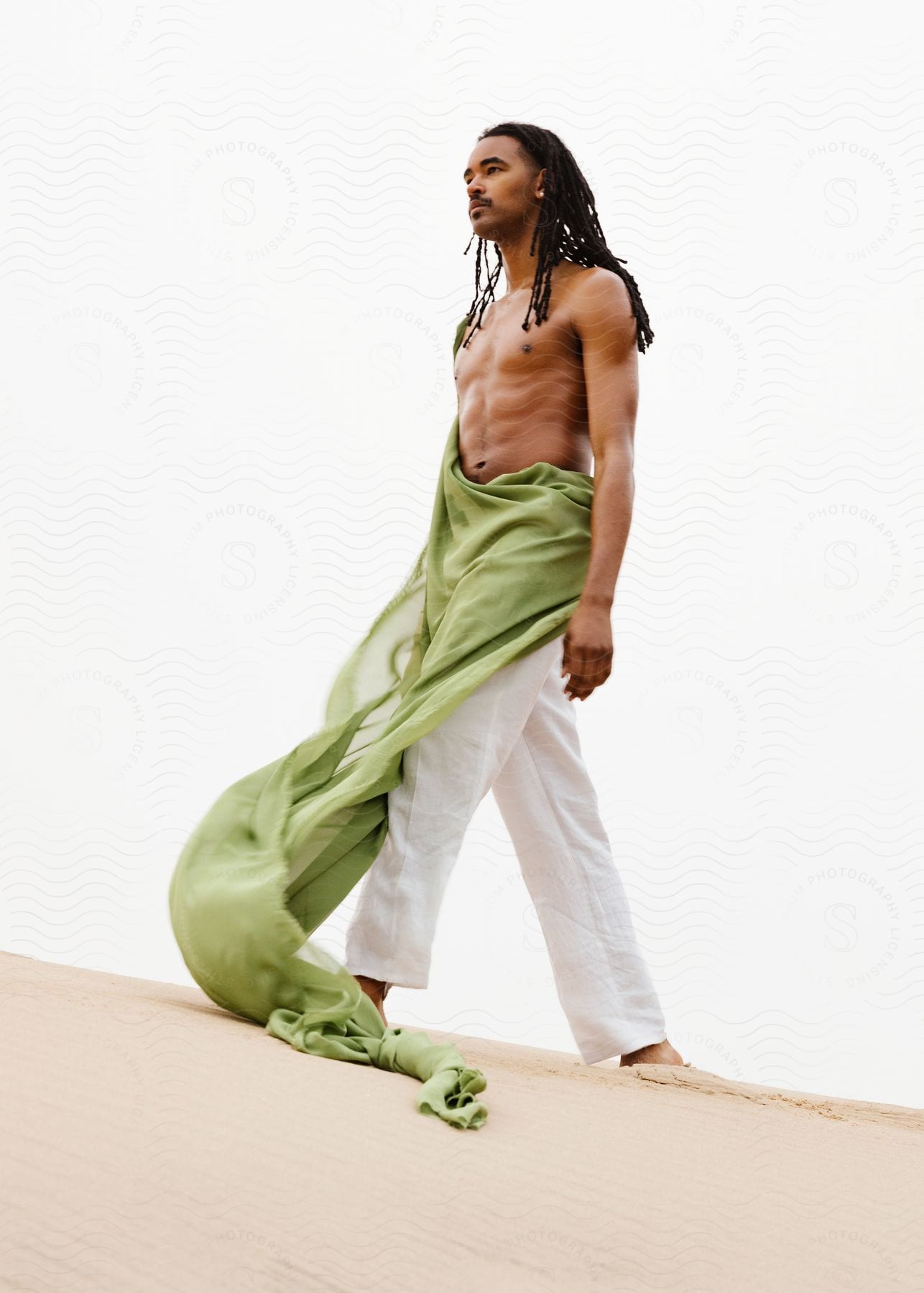 Stock photo of a black man with dreadlocks models a long, green scarf while walking in the sand.