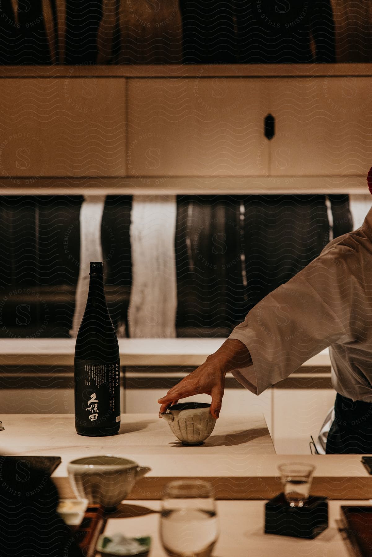 A sushi chef places a small bowl on the counter in front of a sake bottle