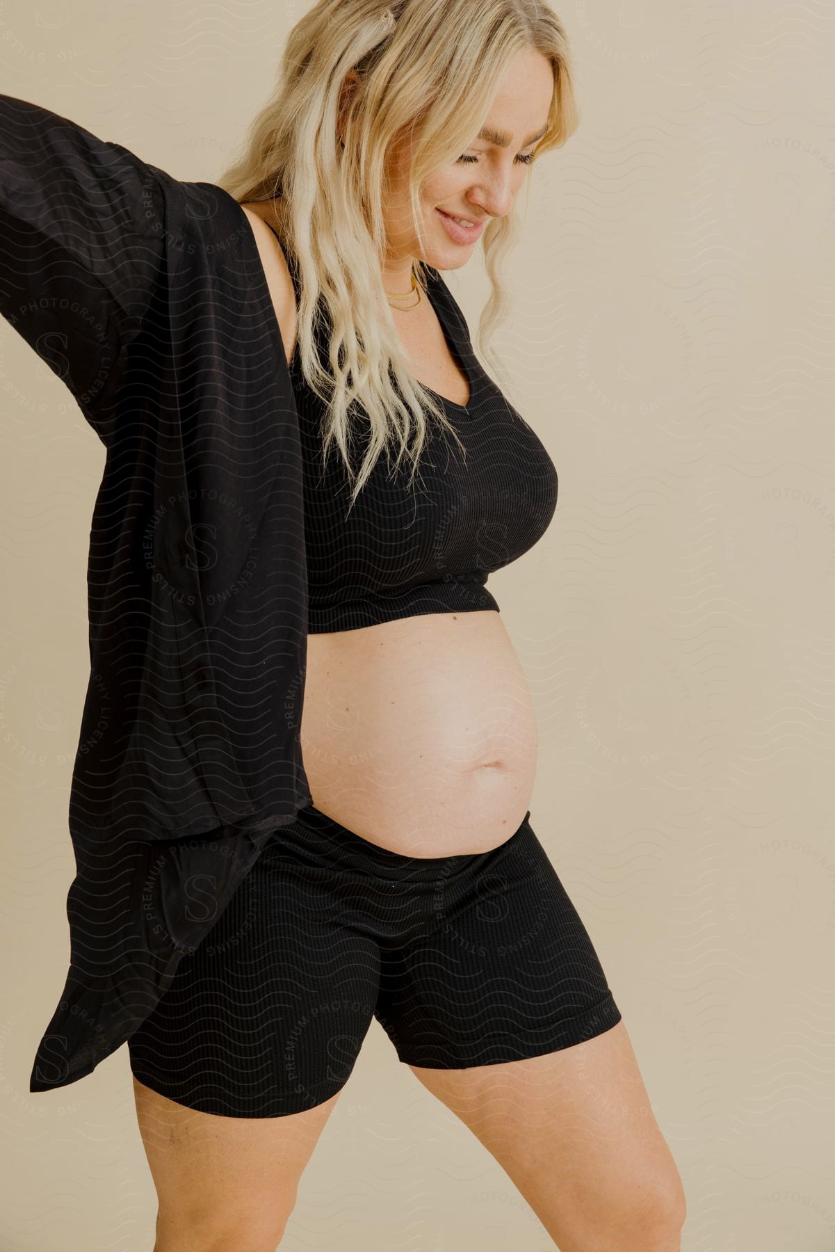 blonde pregnant woman wearing black clothes that reveal her belly standing indoors next to a white wall