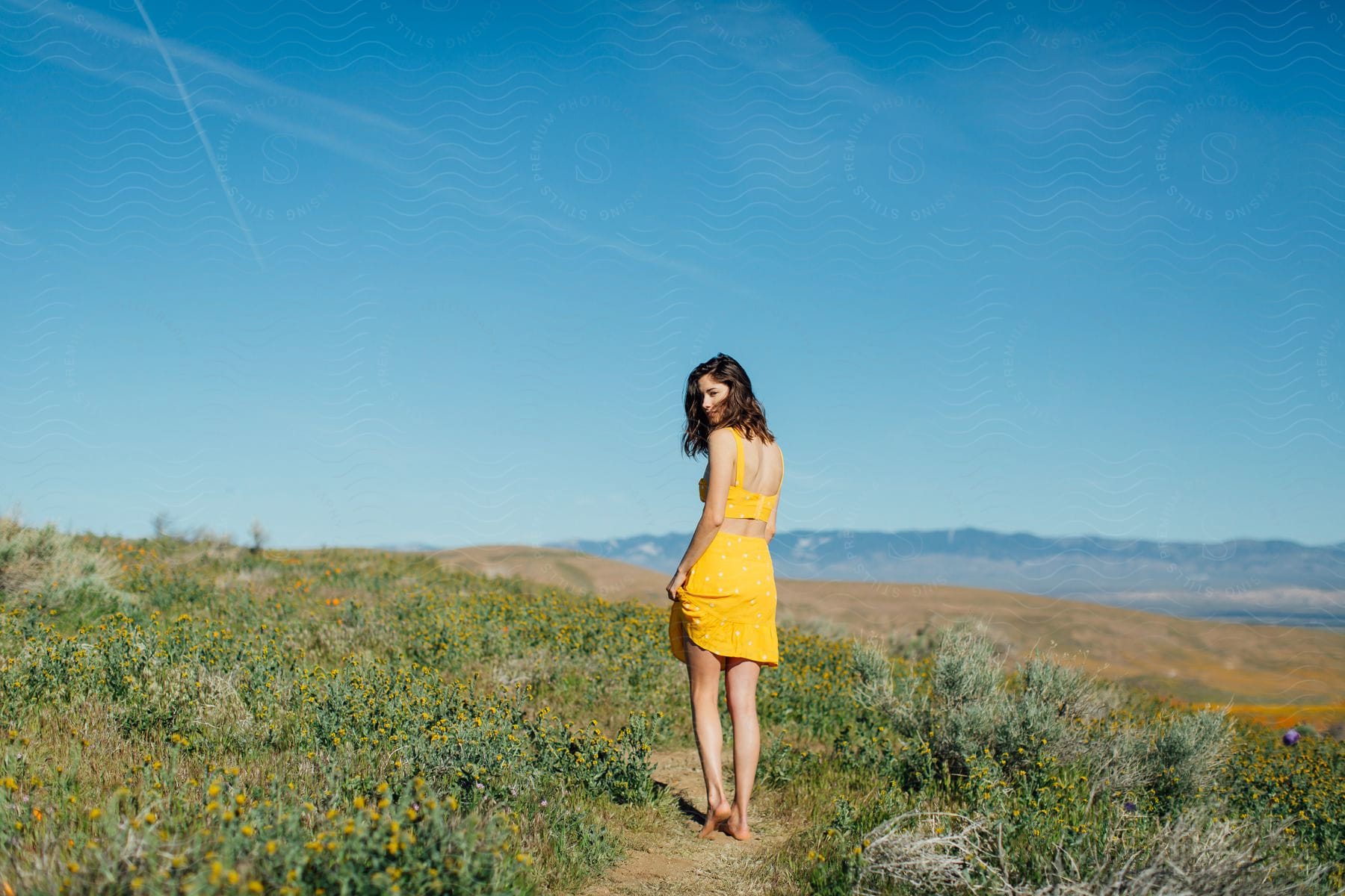 A young woman models a yellow sundress while walking on a hillside trail on a summer day.