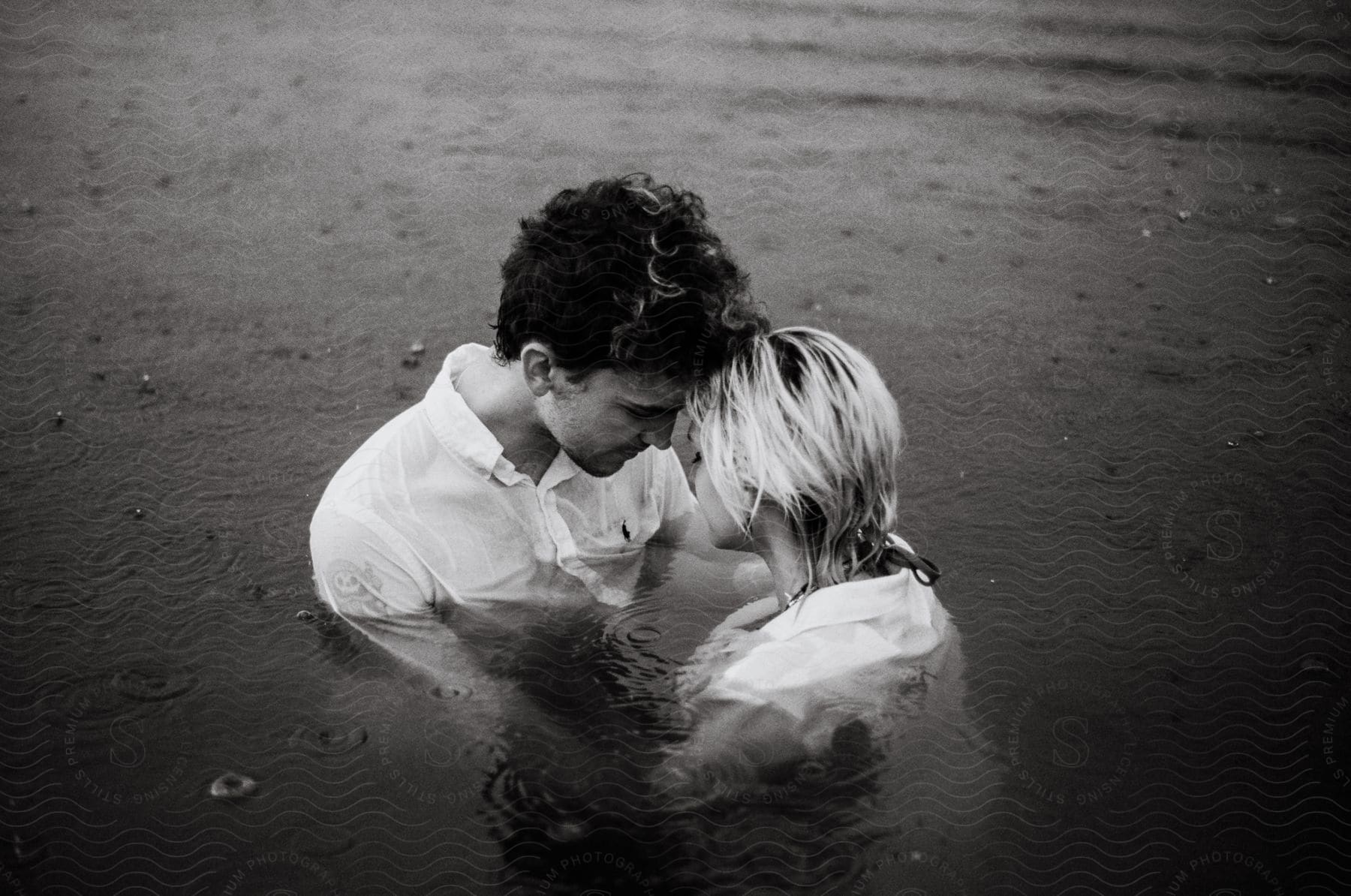 A man and woman in white shirts holding each other in water as it rains.