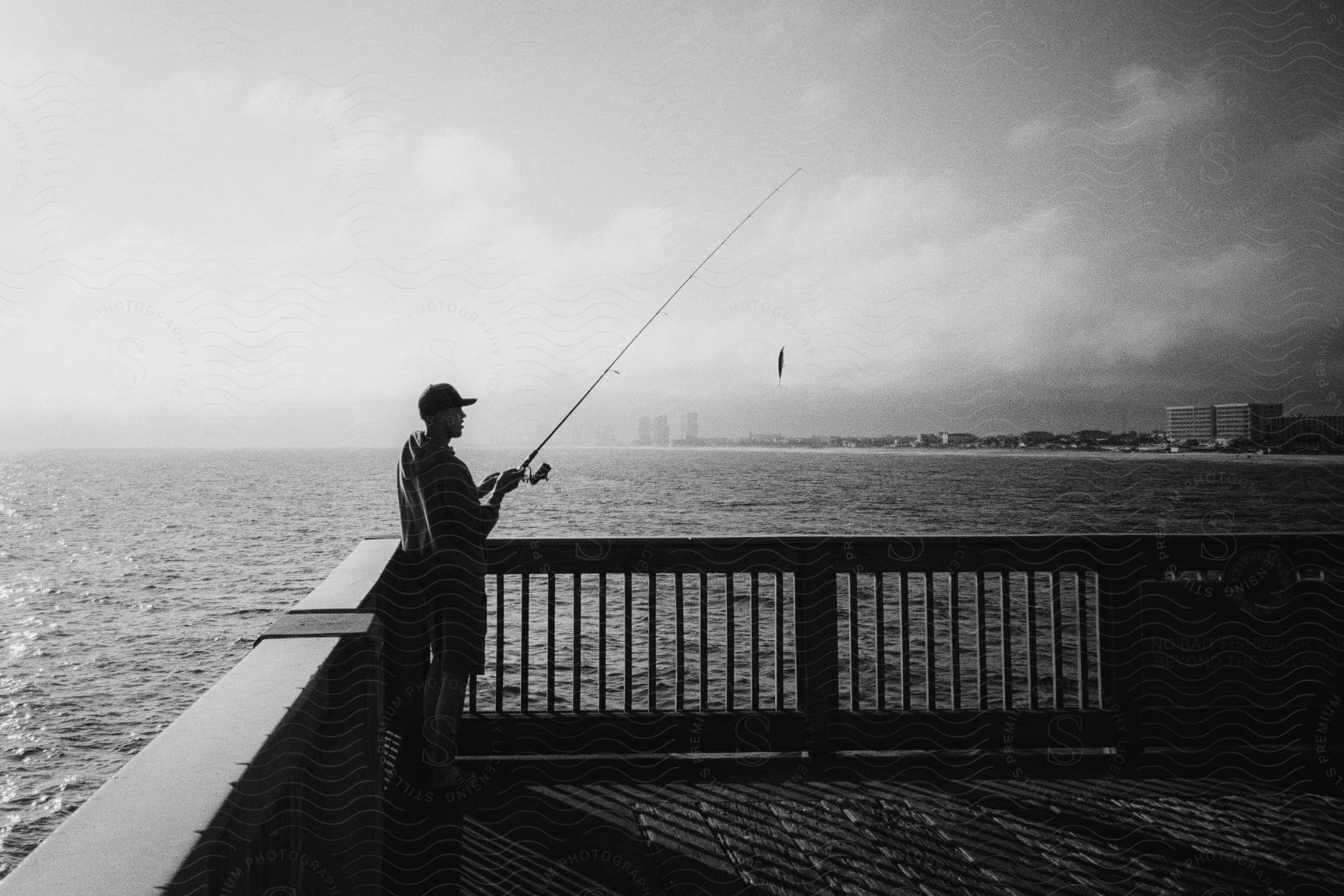 A man stands on a pier while holding up a long fishing rod as the lure dangles from it.