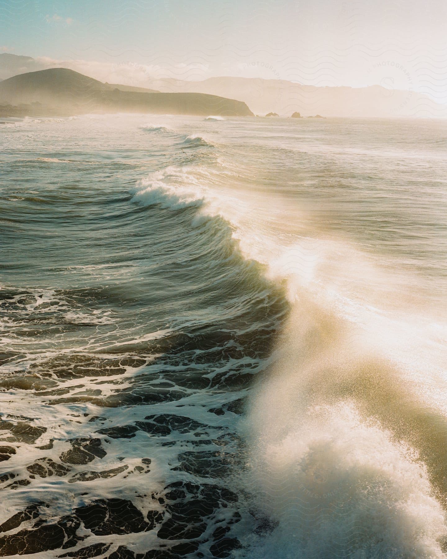 Sun-kissed wave crashes against the coast, framed by majestic mountains.