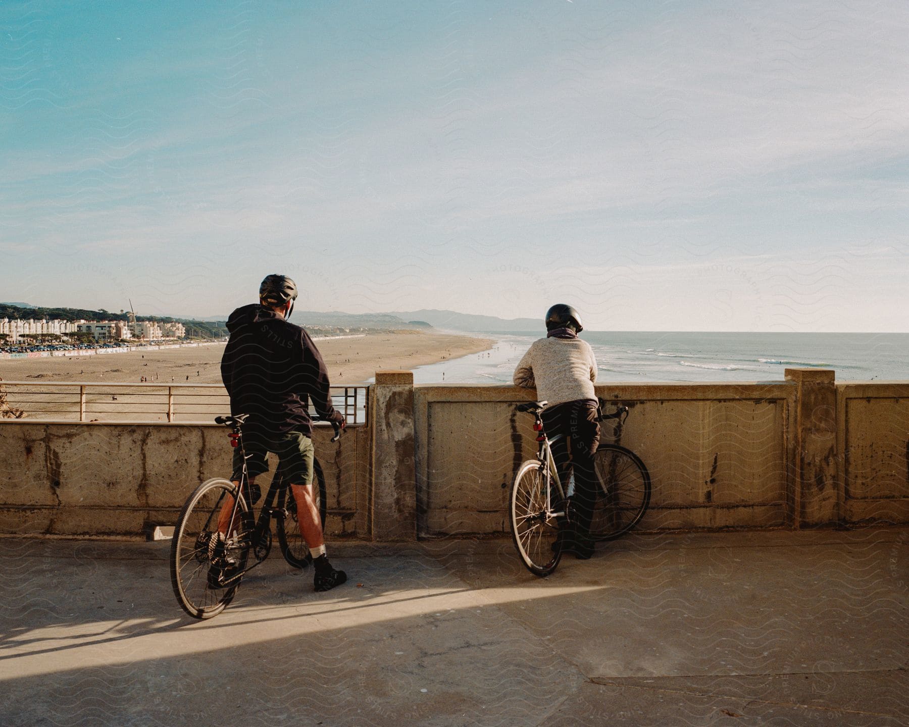 two cyclists outdoors wearing headgear on a bridge next to a beach looking at the ocean during day time