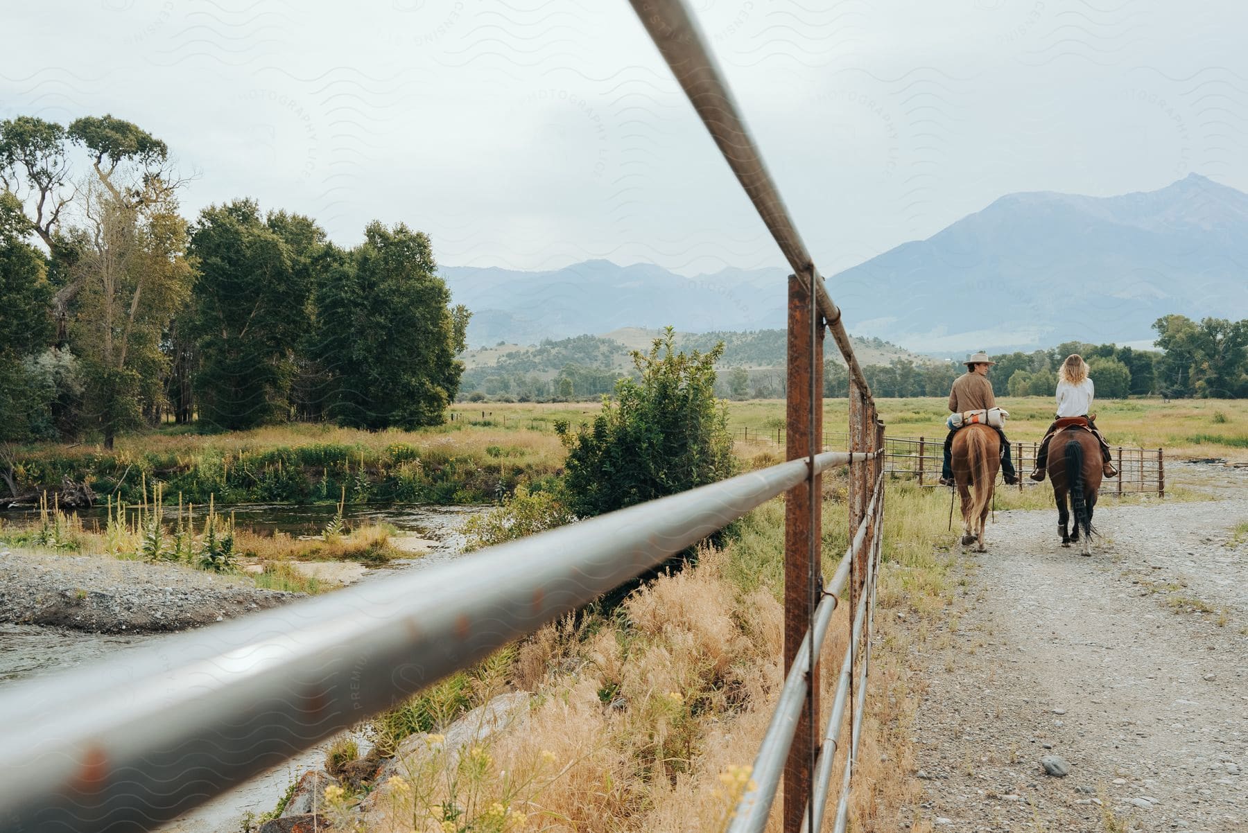 Couple rides horses on dirt road by fence and river, with mountains in the background.