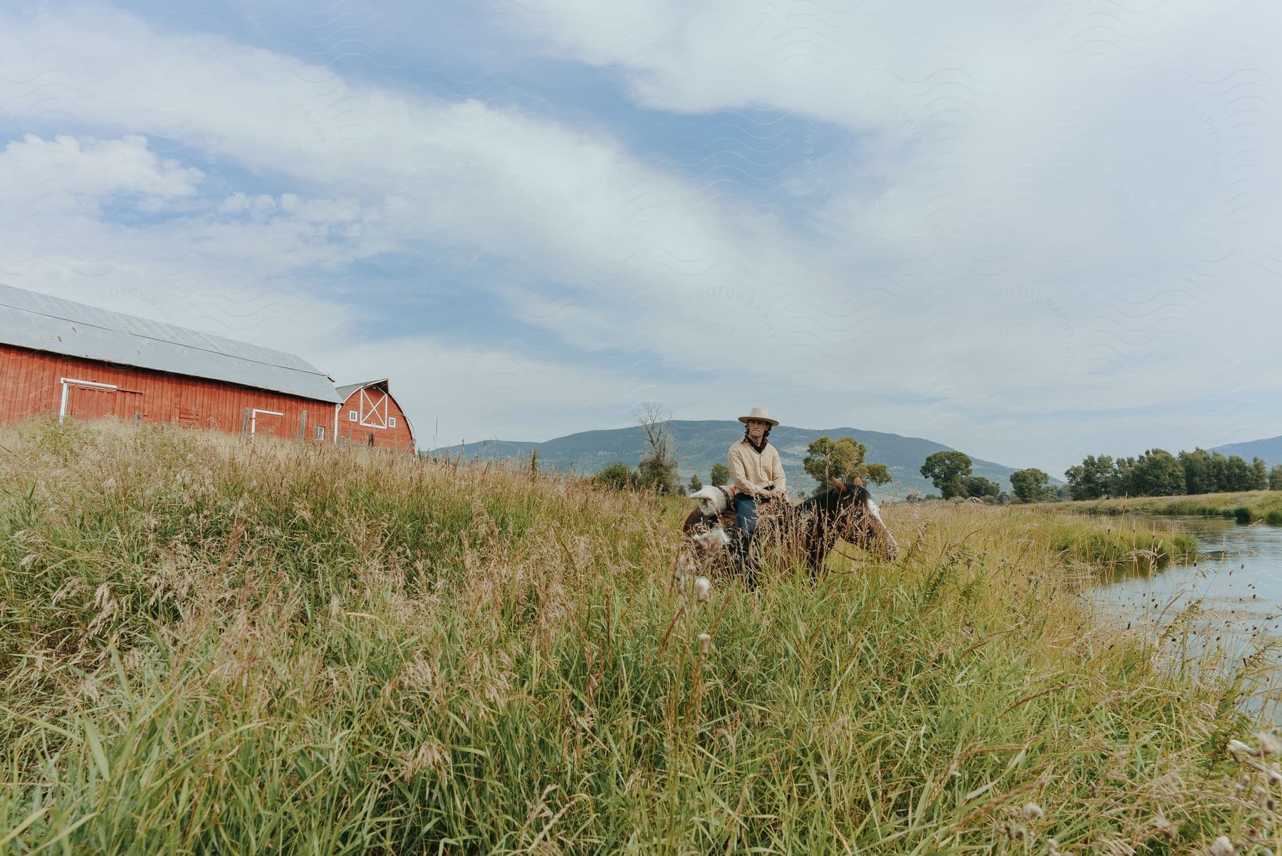 Cowboy sits on horse in field between river and red barn with misty mountains in the background on a cloudy day.