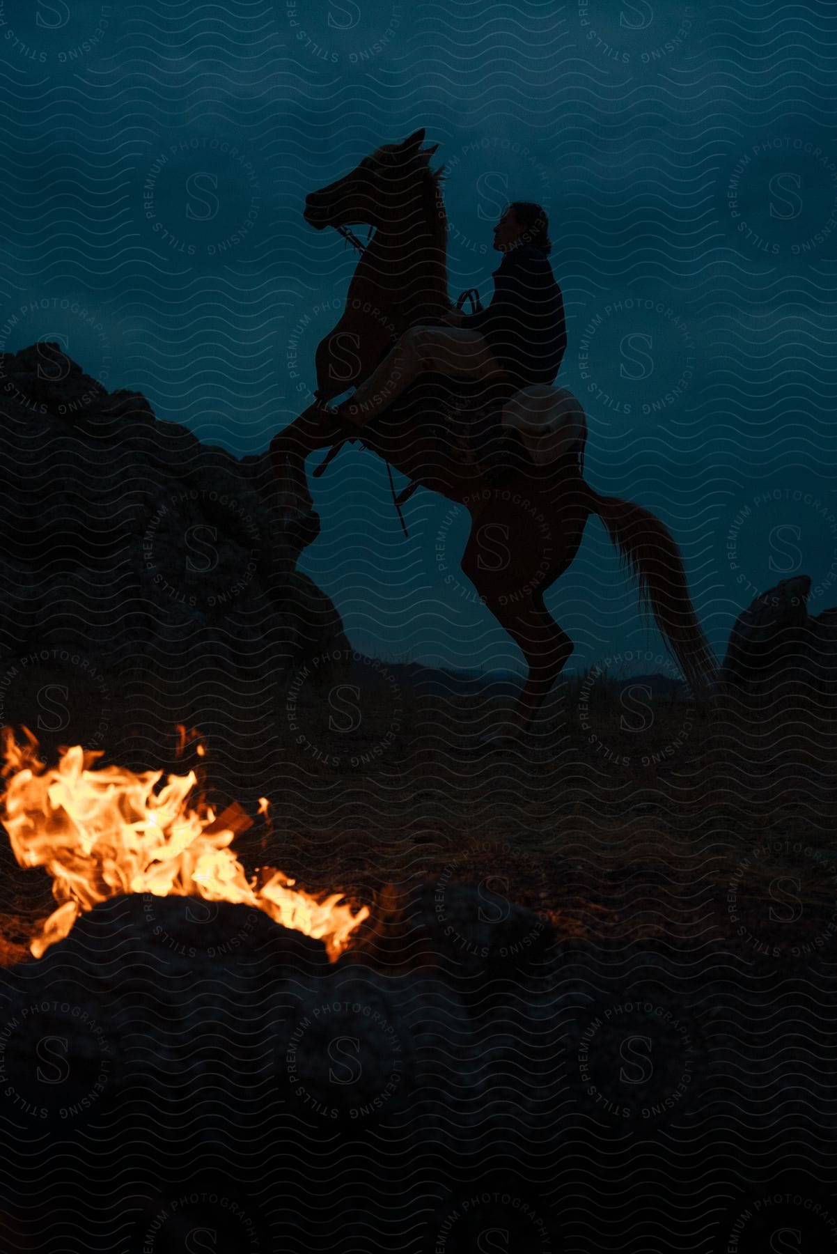 a person sits atop a rearing horse with a campfire in the foreground