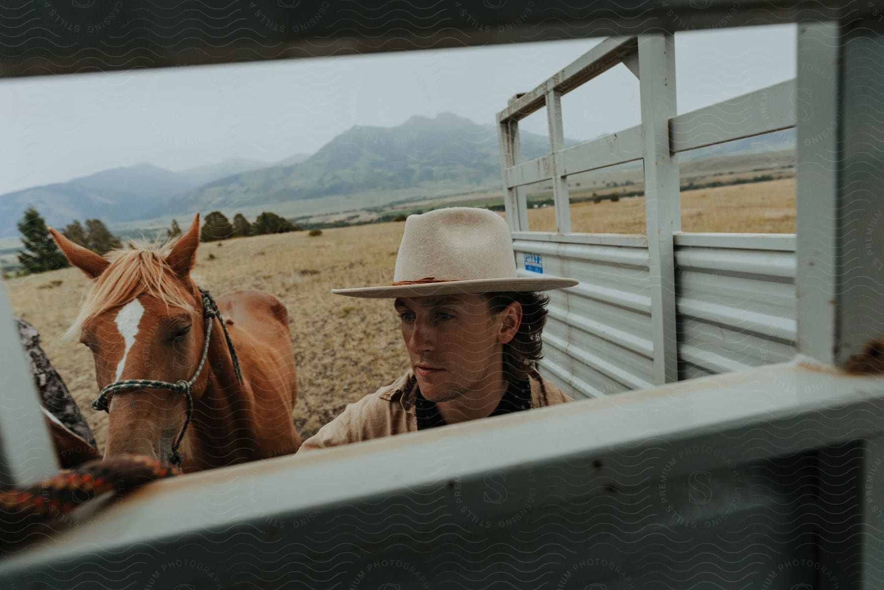 young adult white man outdoors wearing cowboy clothing putting a horse on a stable next to a plain field with vegetation and mountains on the background during day time