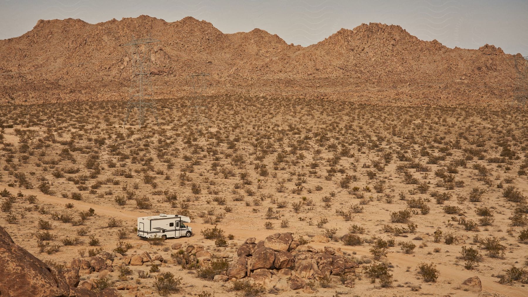 Panorama of a motorhome in the midst of a desert-like terrain with sparse vegetation and rocky mountains in the background.