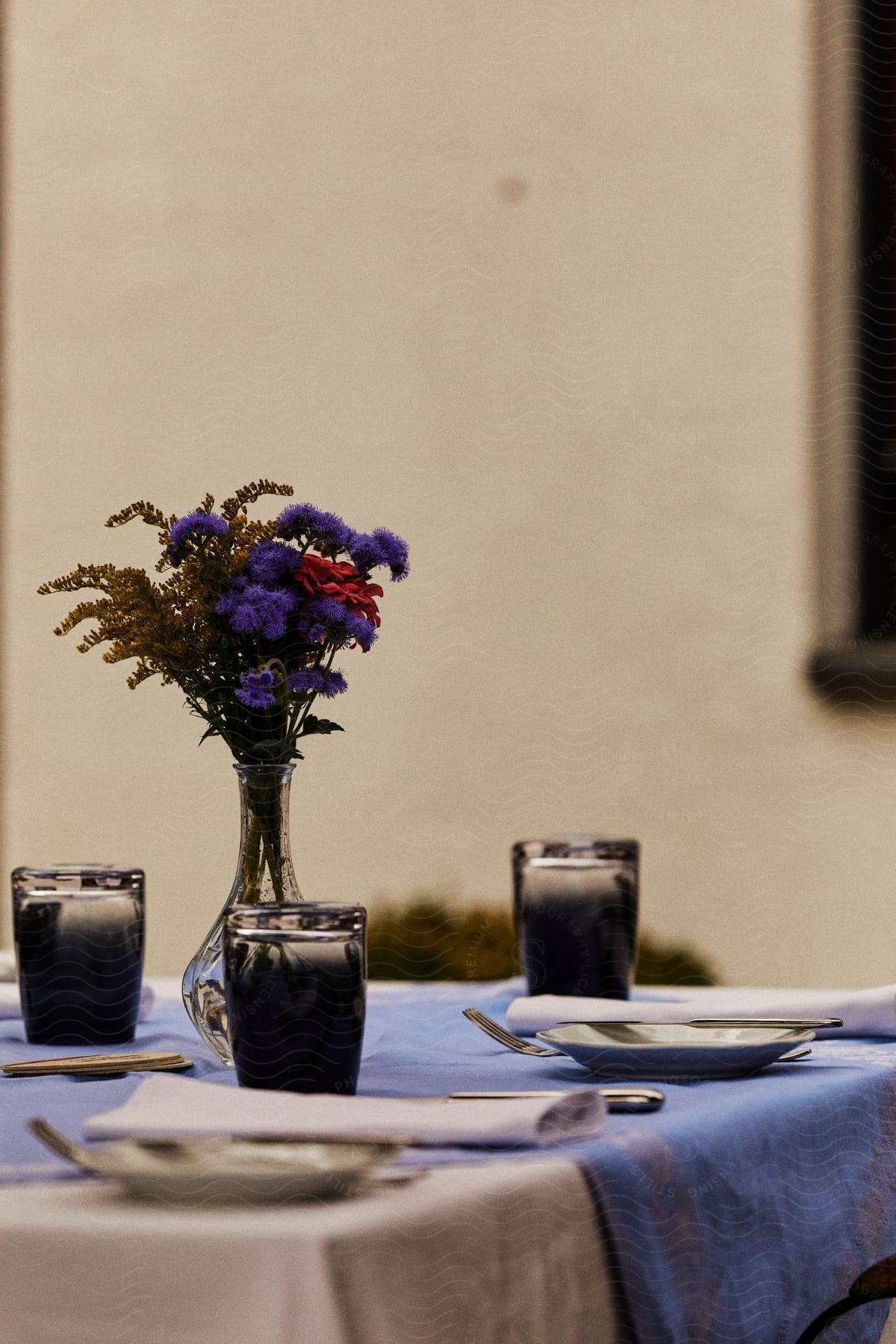 A dining table where there are glasses, cutlery and plates and a vase with purple red and yellow flowers