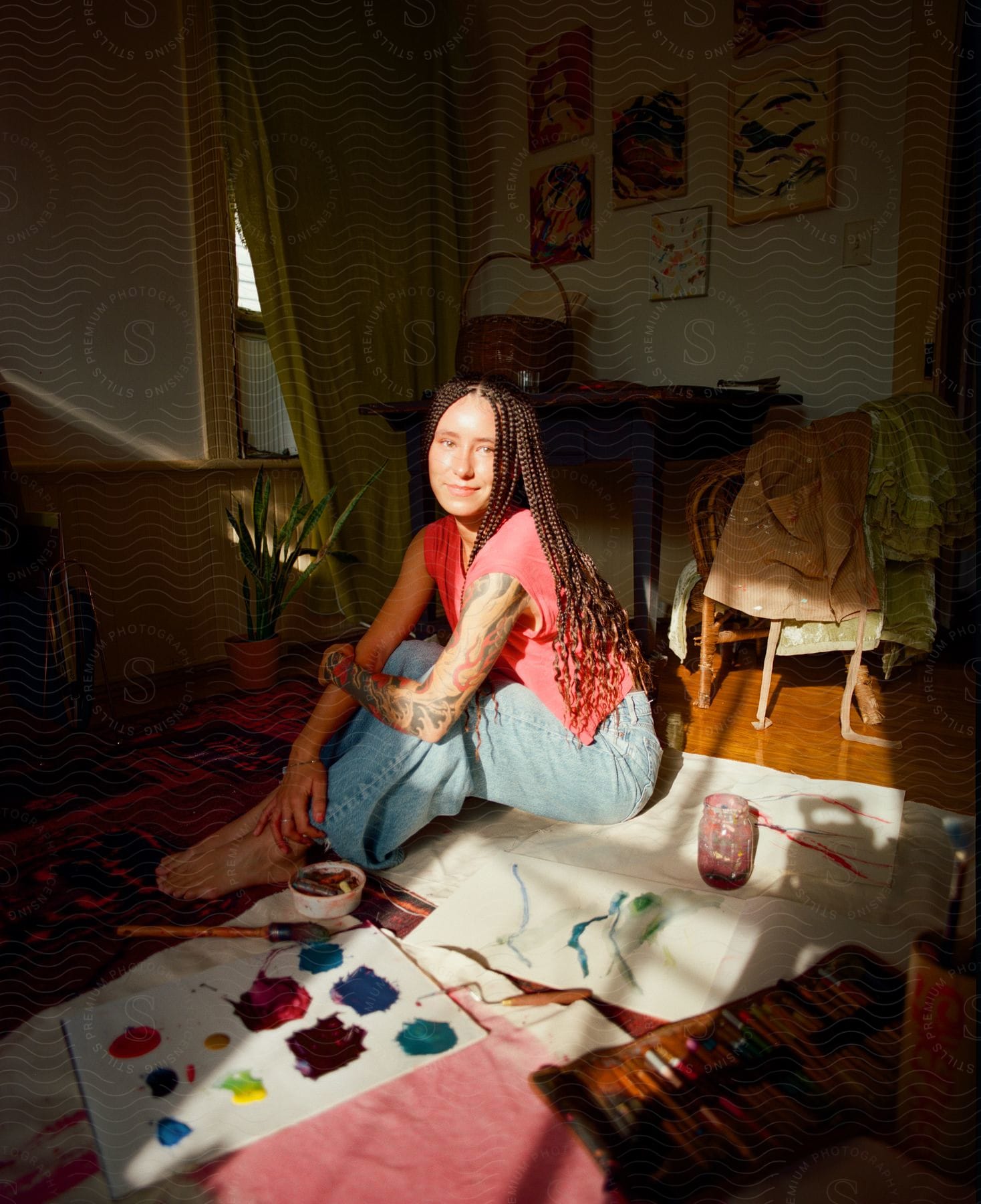 Woman with braided hair sitting on floor with paint and brushes, artwork on walls in background, natural light from window.