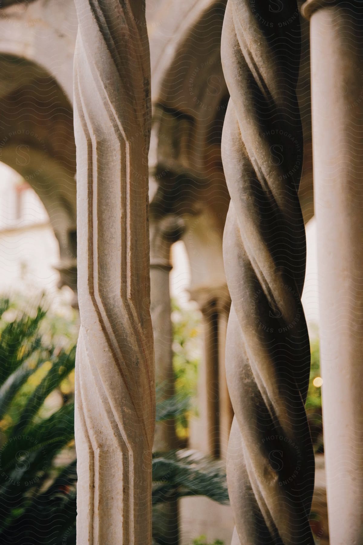 Roman style columns of a building two different spiral designs with archways and plants in the background during the day.