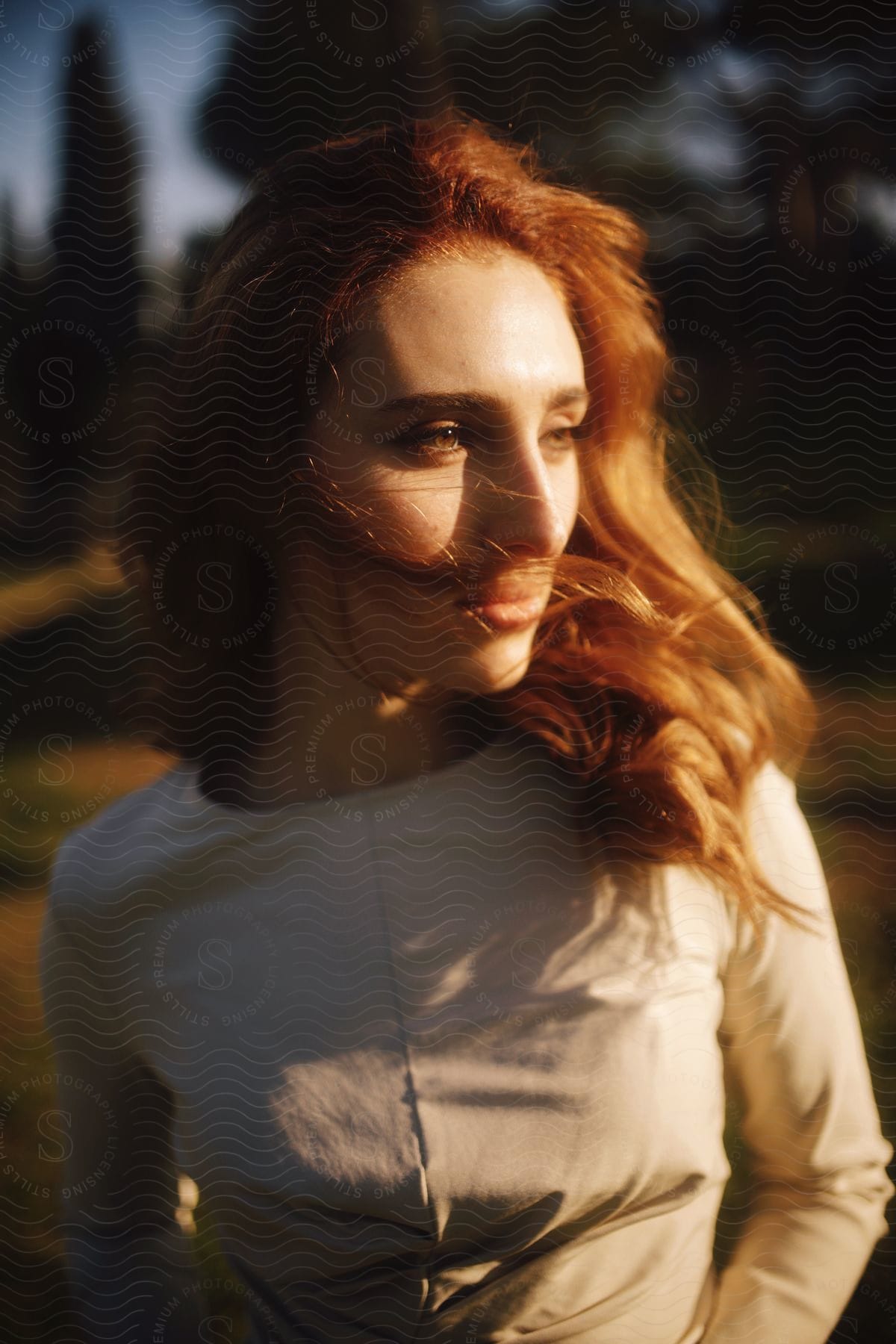 Sunlight shines onto face of redheaded woman standing in field in fashionable attire.