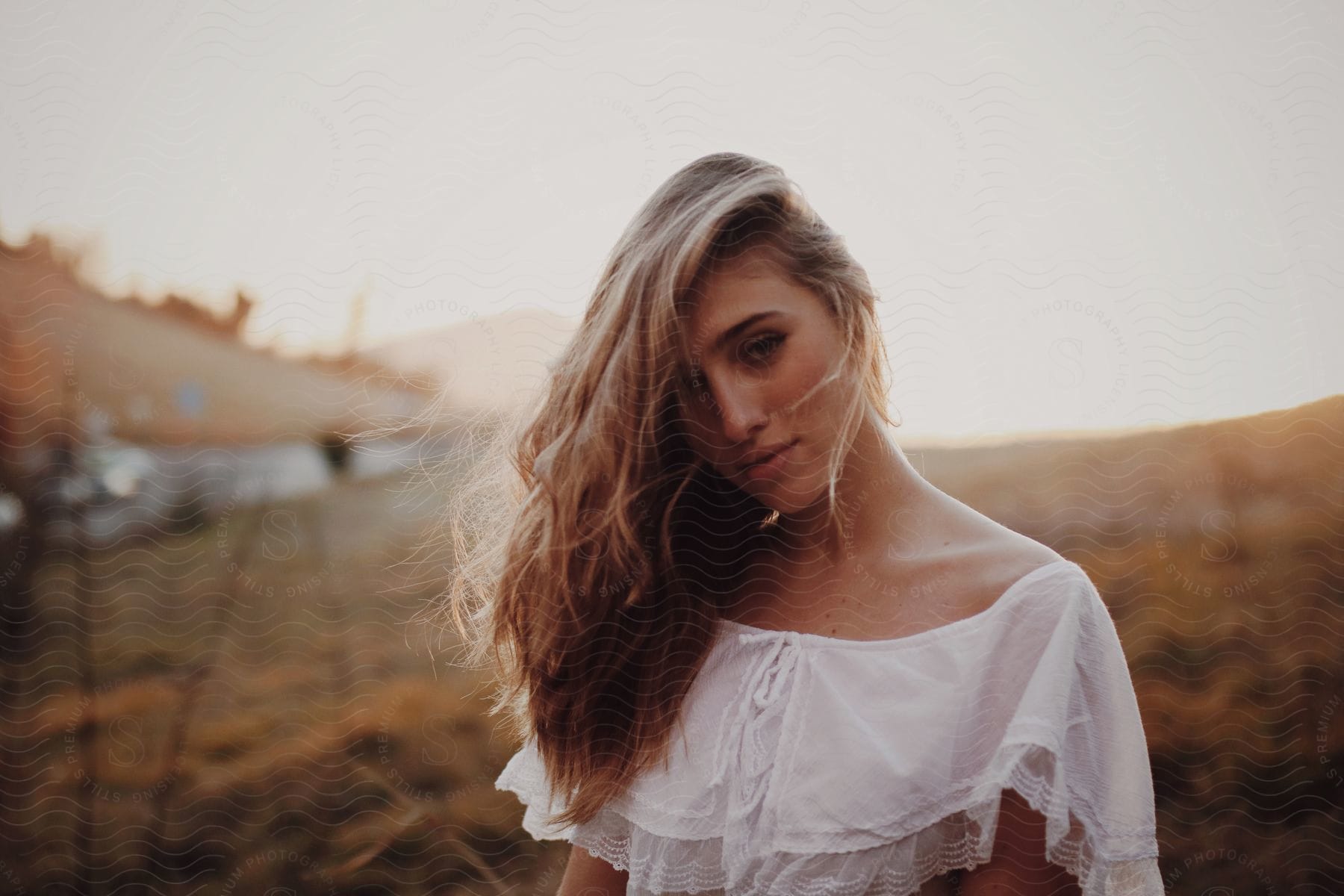 A woman wearing a romantic white top posing on a hillside with her long hair brushed over to one side