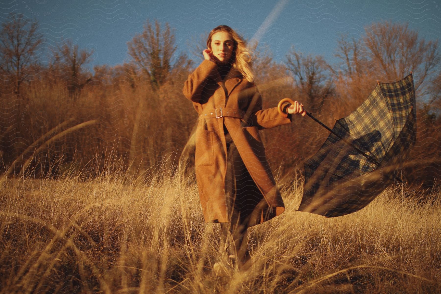 Blonde model in a field posing with a long brown coat and an open umbrella.