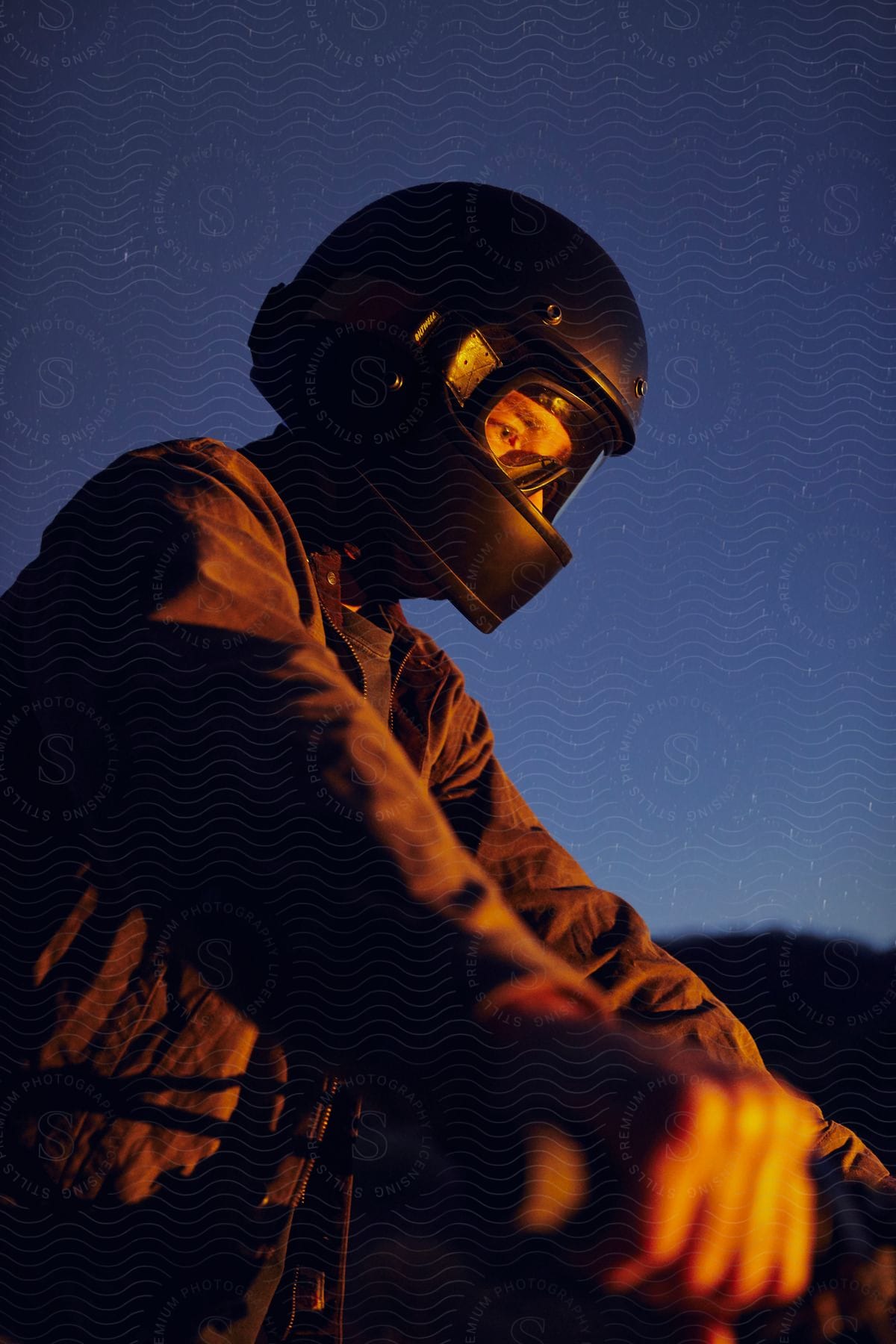 A motorcyclist wearing a full-face helmet is captured in a contemplative moment, illuminated by warm light against the evening sky.