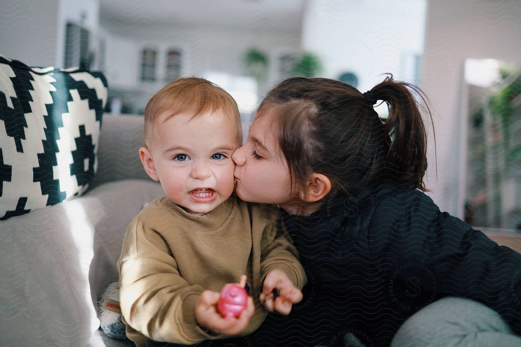 Sweet sister plants a kiss on baby brother's cheek, cuddling on a gray couch.