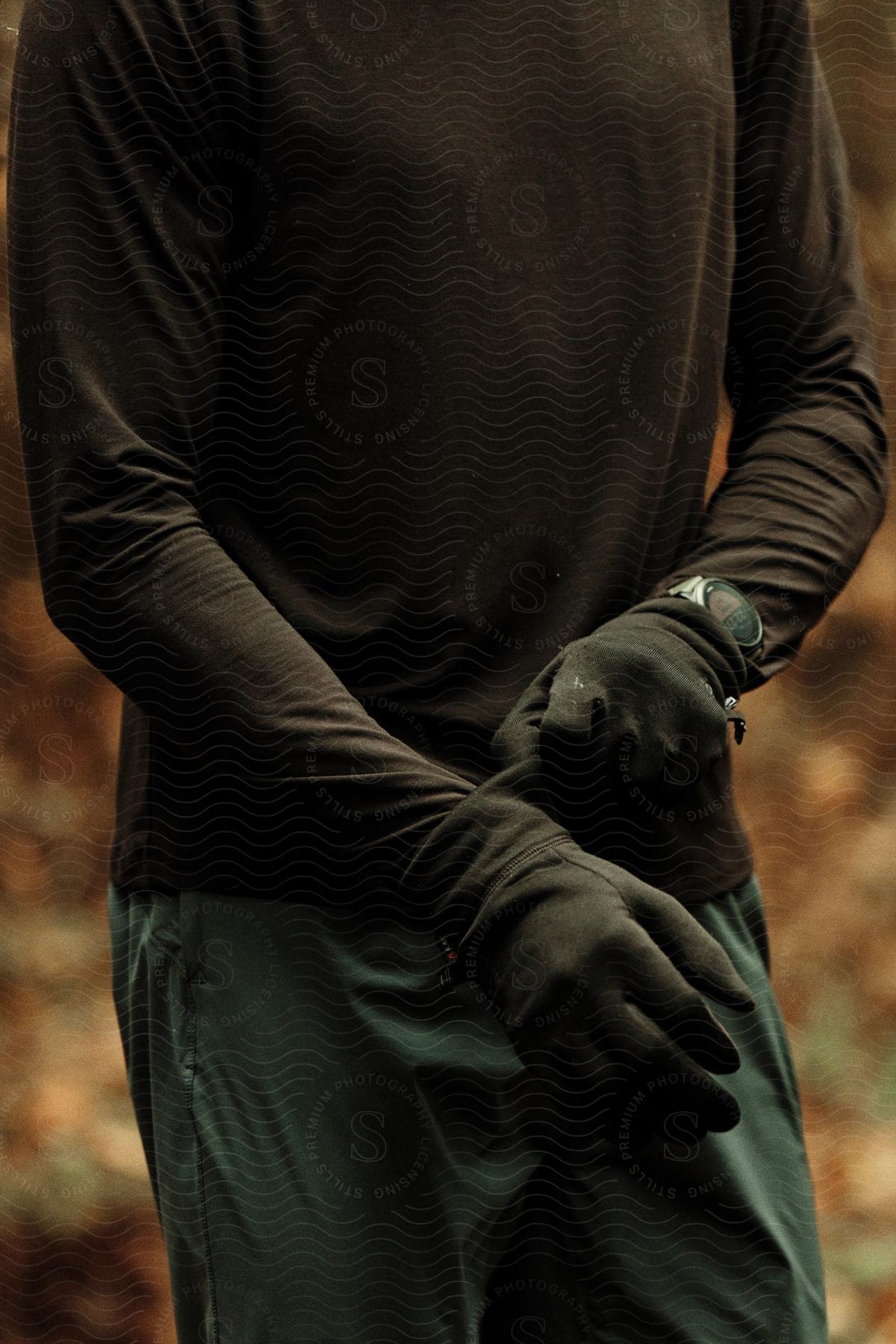 A man wearing a black long sleeve shirt, green pants, and black gloves with a fitness tracker on his wrist stands against a backdrop of woods with fallen orange leaves in the blurry background.