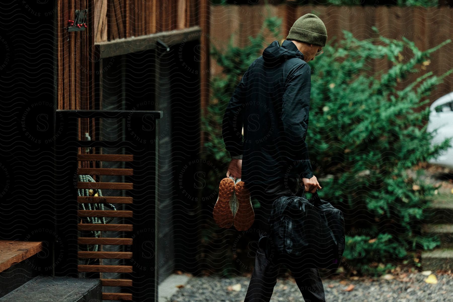 A man in a windbreaker walks out of a house with a bag of luggage in one hand and orange shoes in the other, heading towards a white car parked by closed garage doors.