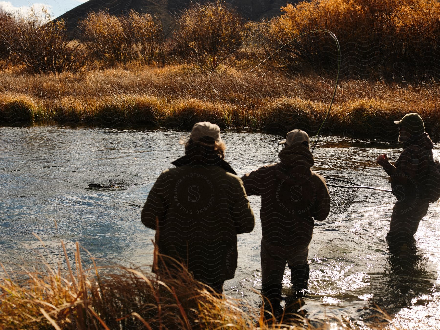 Three bundled-up friends fish in a grassy riverbank, one reeling in a fish with a net and rod.