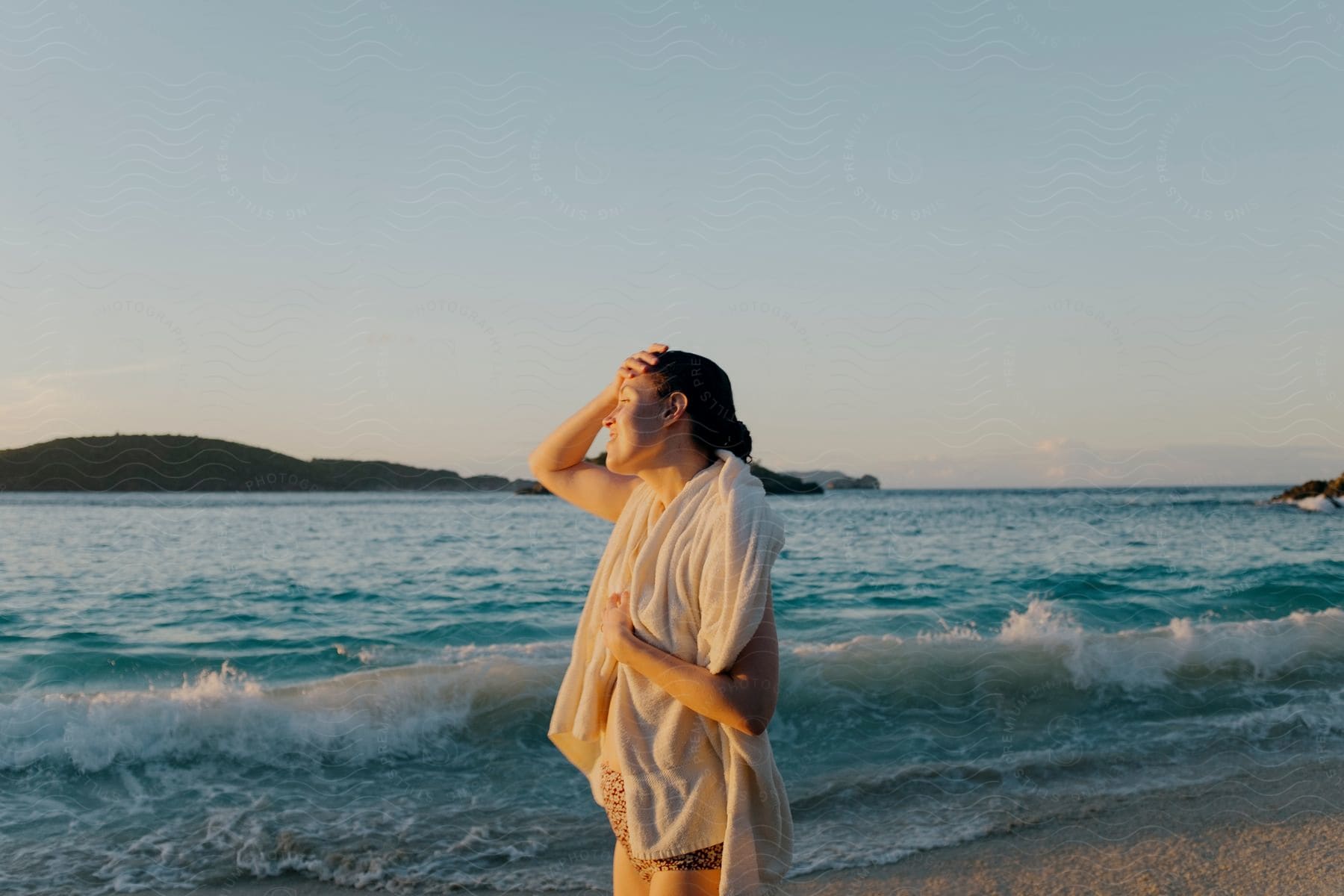 A woman stands on a beach with a white towel around her neck, beside the ocean with waves and blue water, her eyes closed, hair in a bun, with a sunset and an island on the horizon.