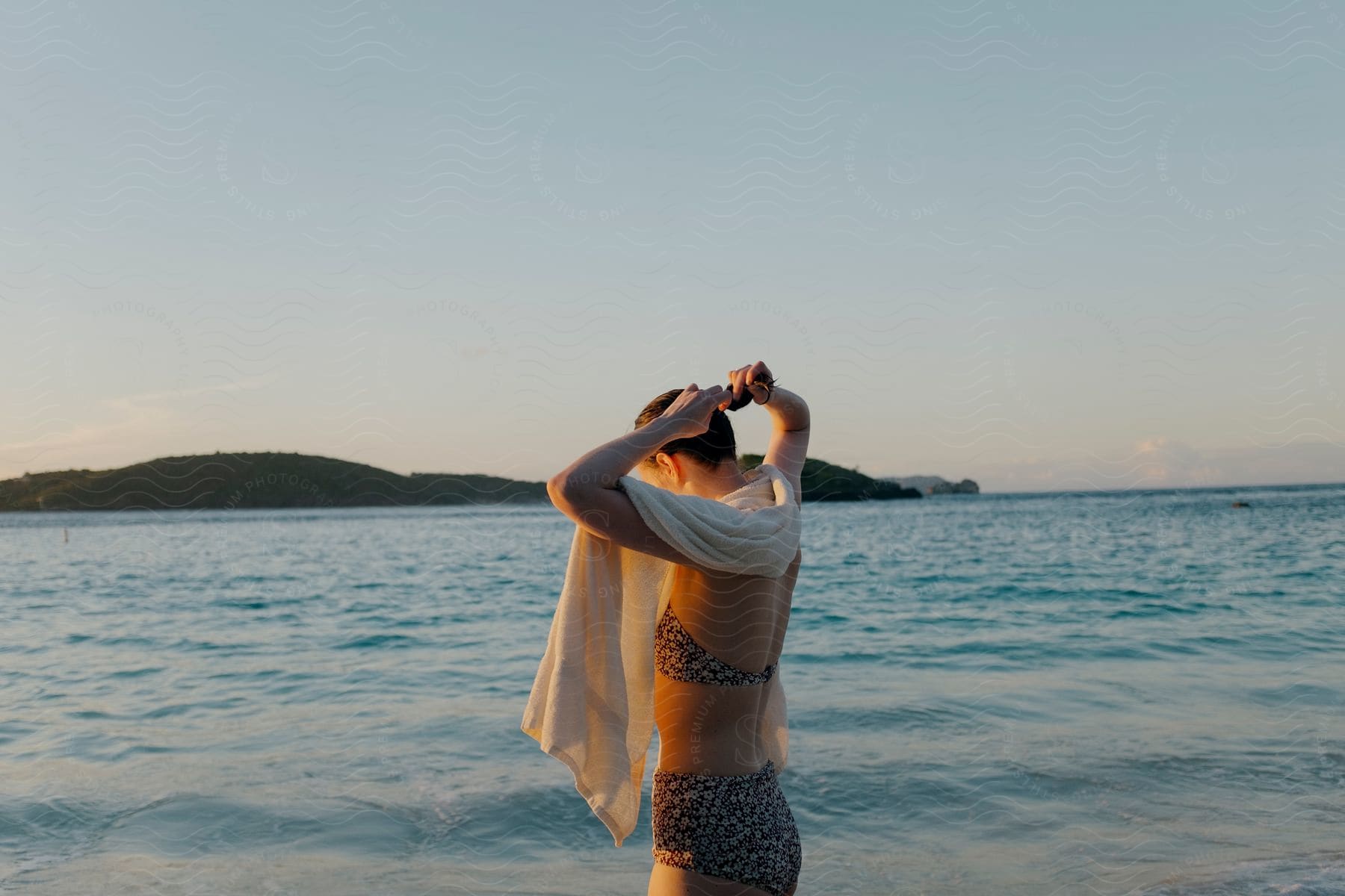 a woman using a towel on the beach while tying her hair and wearing a bikini