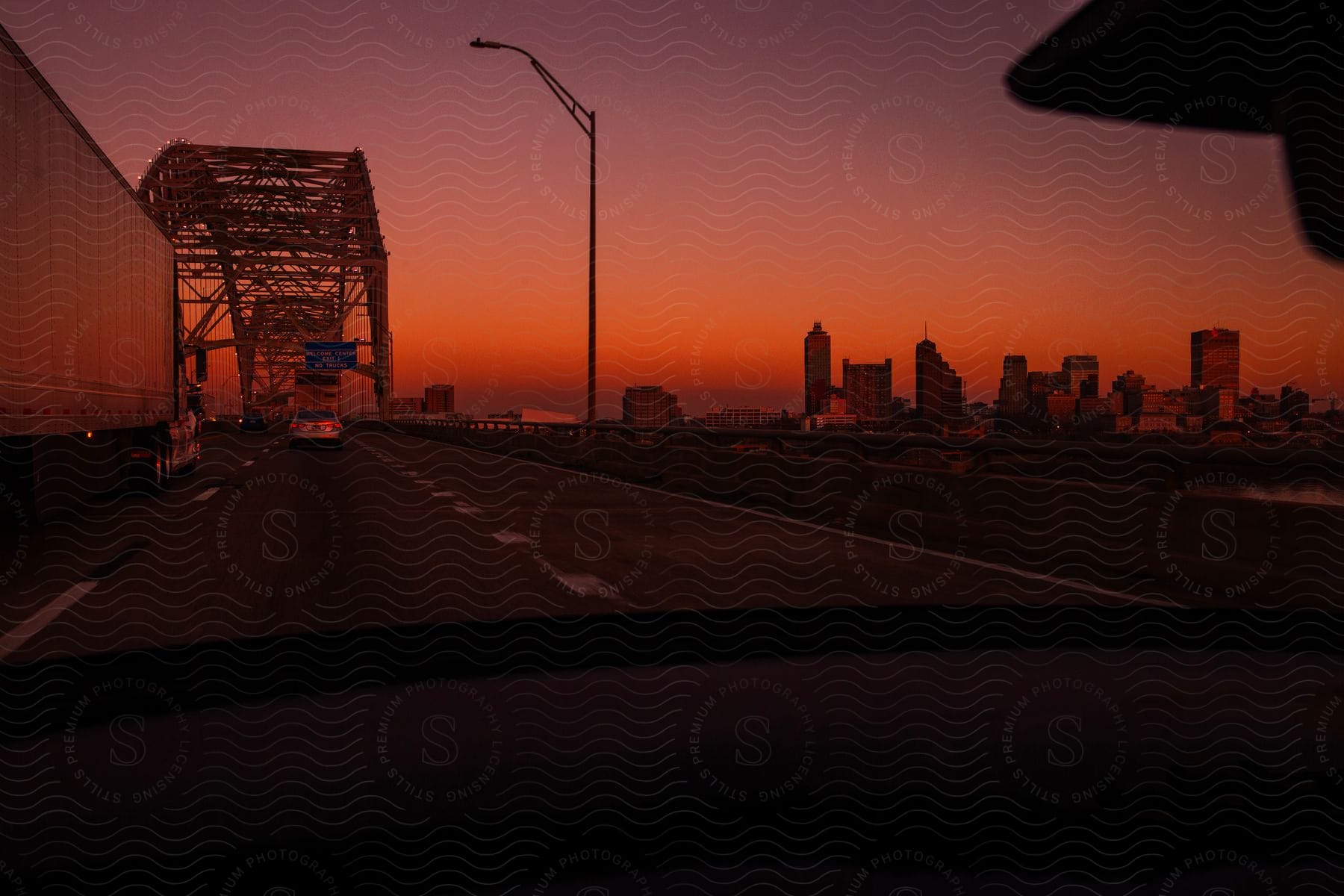 Spooky orange sunset casts an eerie glow on the city skyline seen from a car on a steel bridge with freight trucks rumbling by.