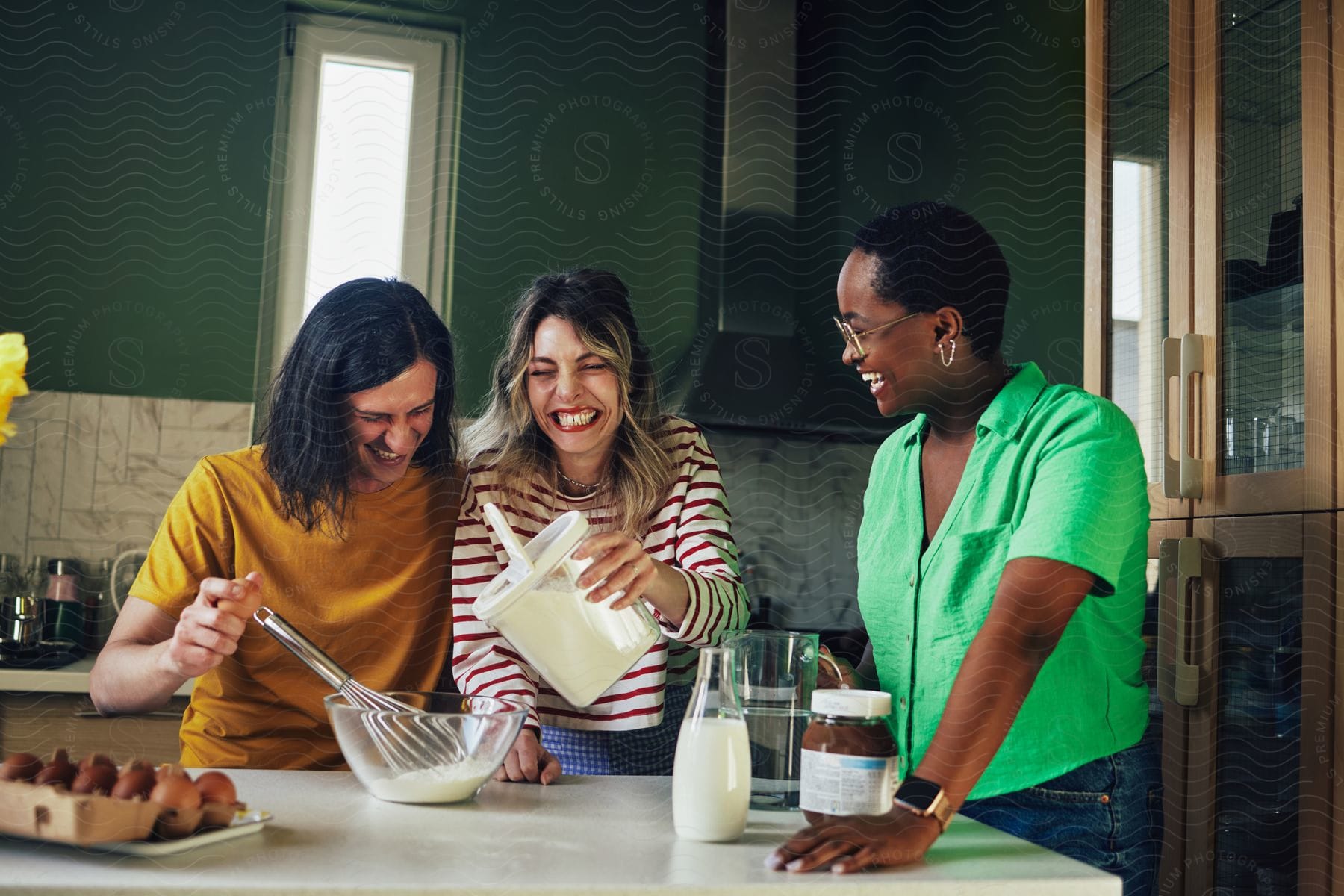 Three friends are smiling in a kitchen while pouring ingredients into a mixing bowl.