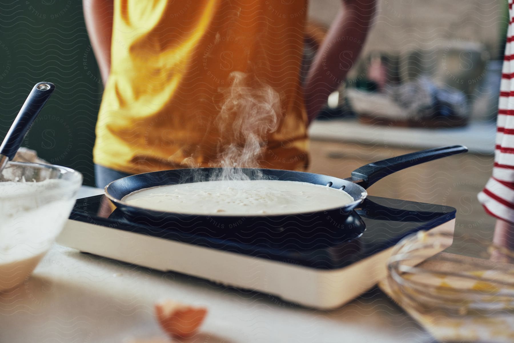 People cook batter in shallow skillet placed on portable stove in kitchen.