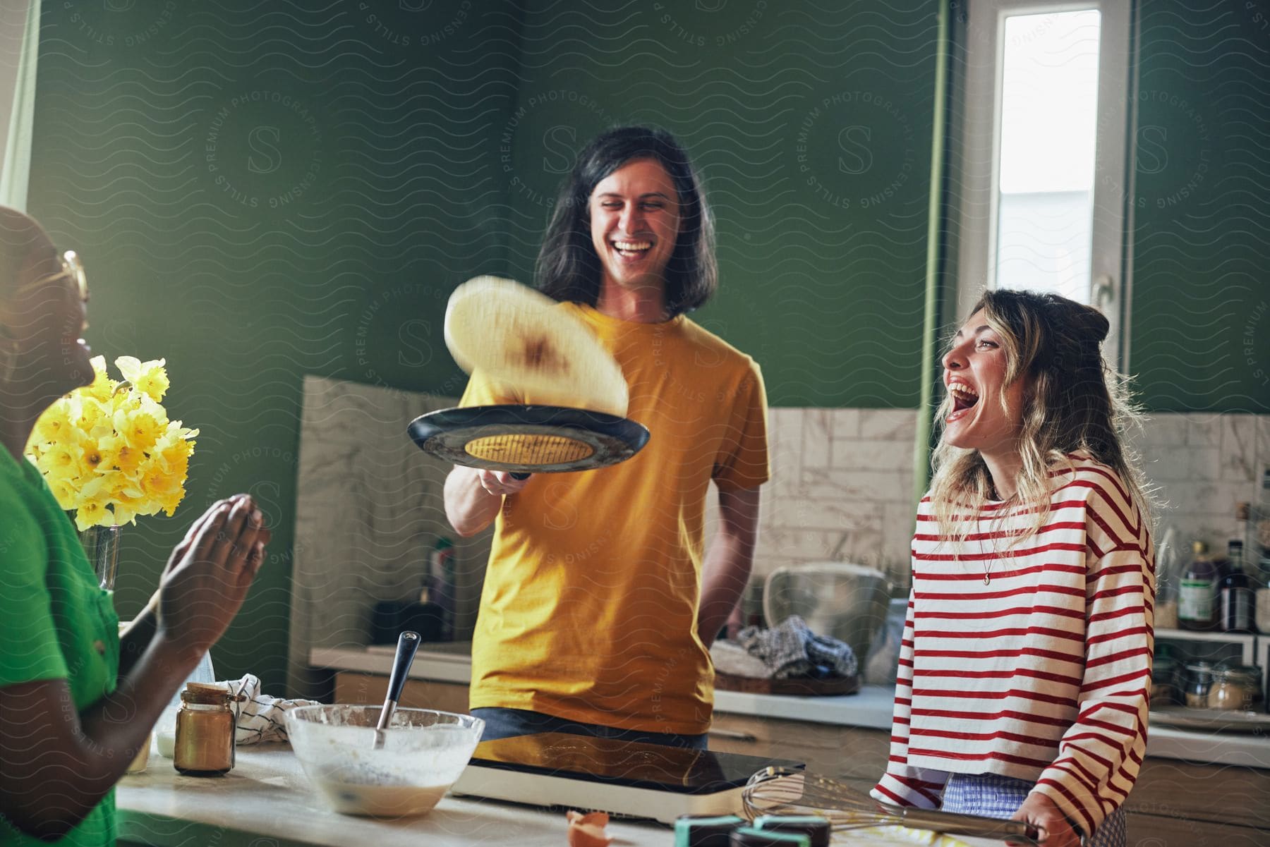 Two people stand in the kitchen laughing as they flip food in a pan and another person watches